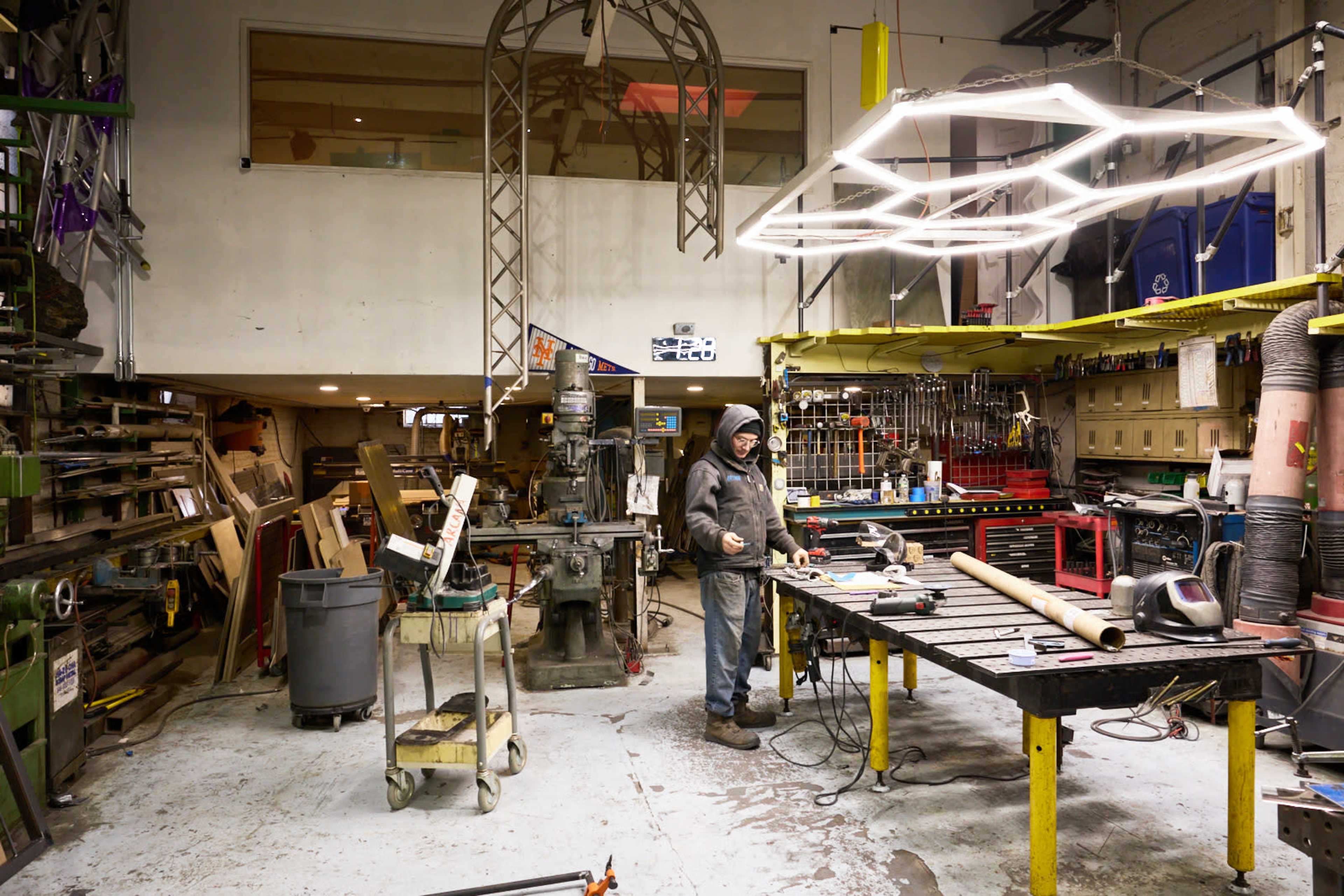 A person in a hoodie works at a metal table in a cluttered workshop filled with tools, machinery, and various materials.