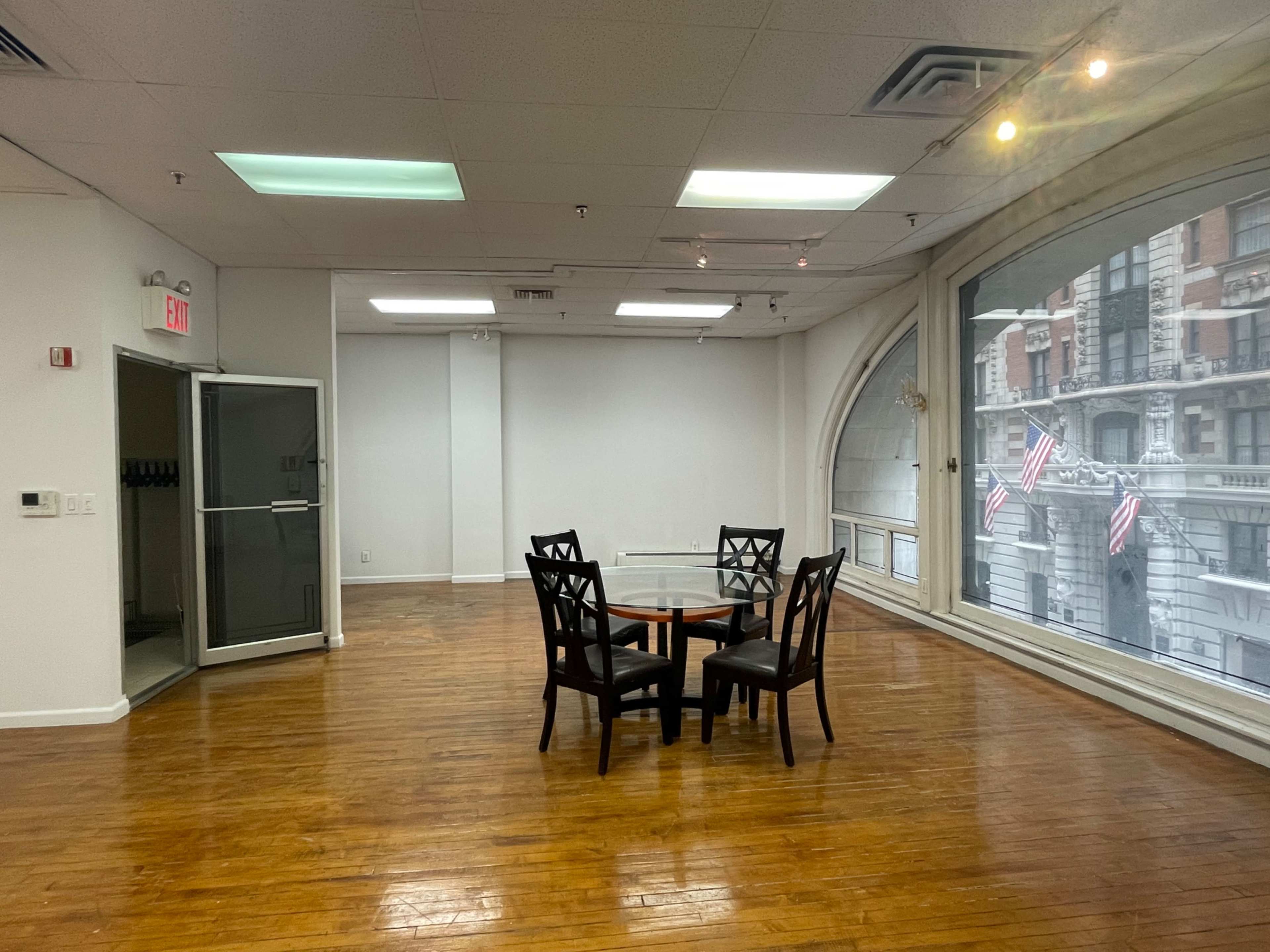 A vacant room with hardwood floors features a round dining table surrounded by four black chairs, illuminated by natural light from large windows displaying an outdoor view with flagpoles.