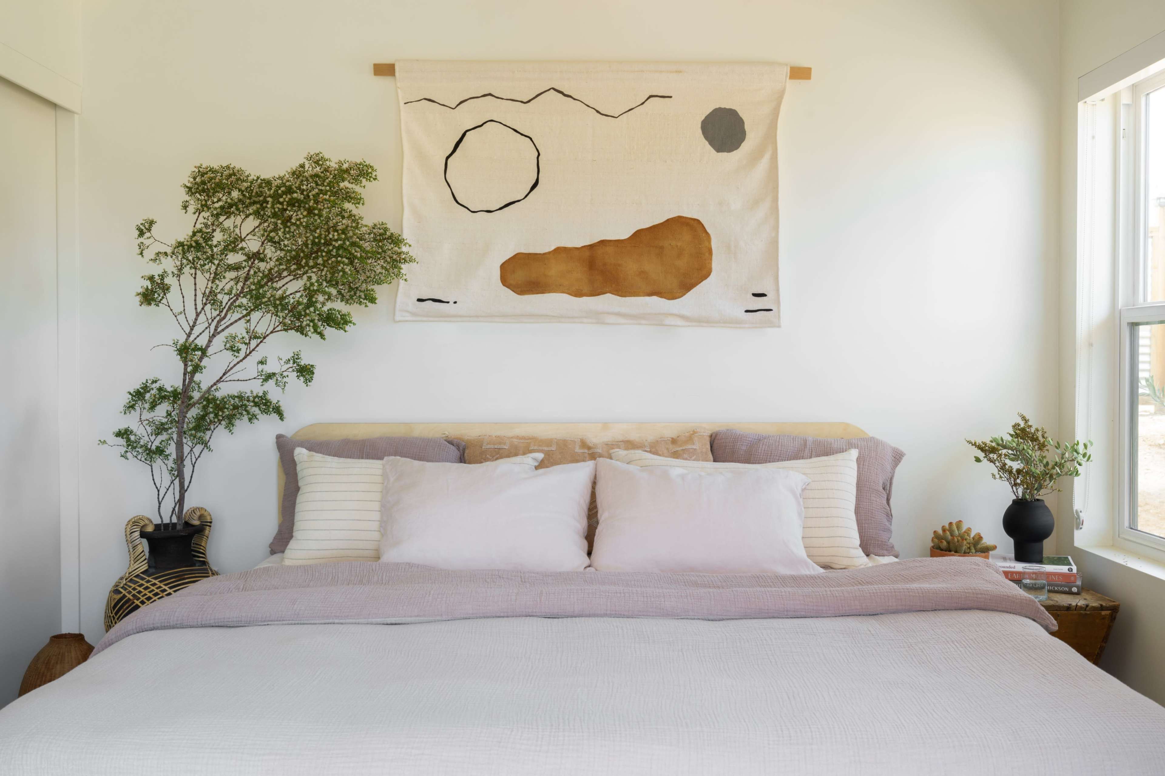 A neatly arranged bedroom features a wooden bed with pillows, a woven wall hanging above, and potted plants by the window.