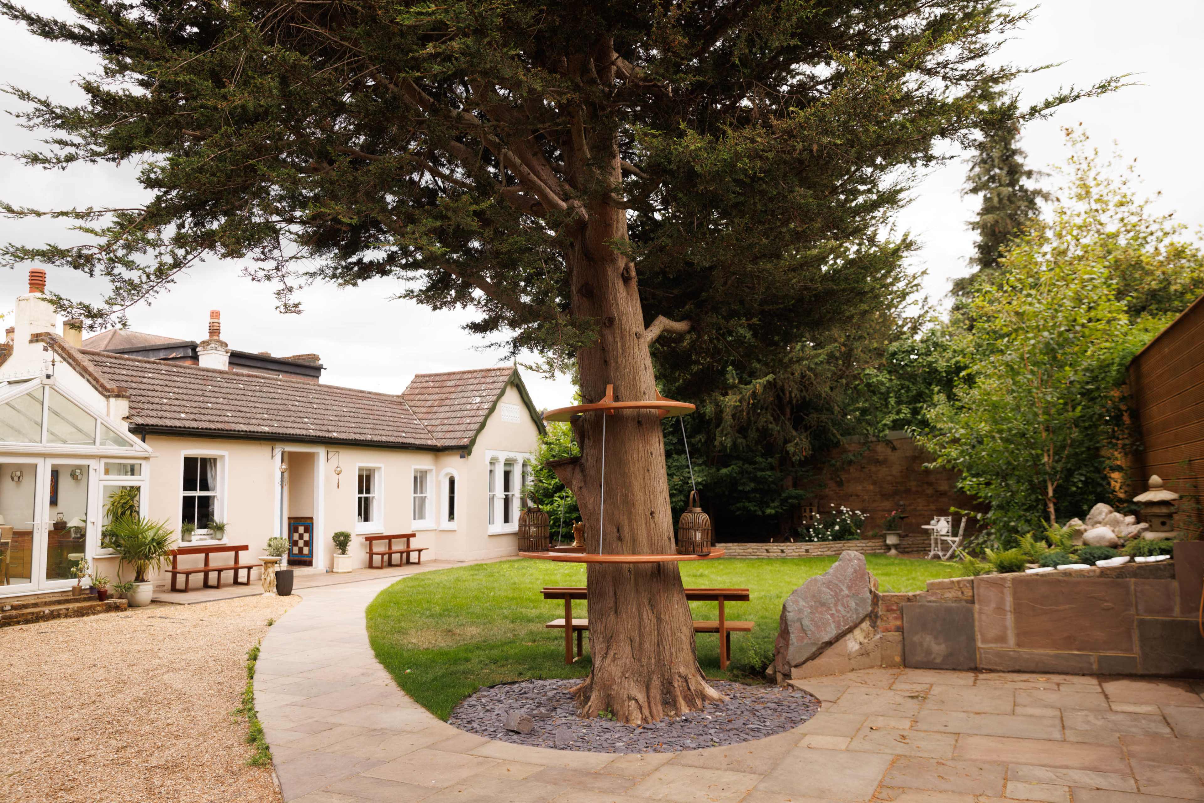 A large tree with wooden shelves stands in a grassy yard beside a pathway that leads to a house.
