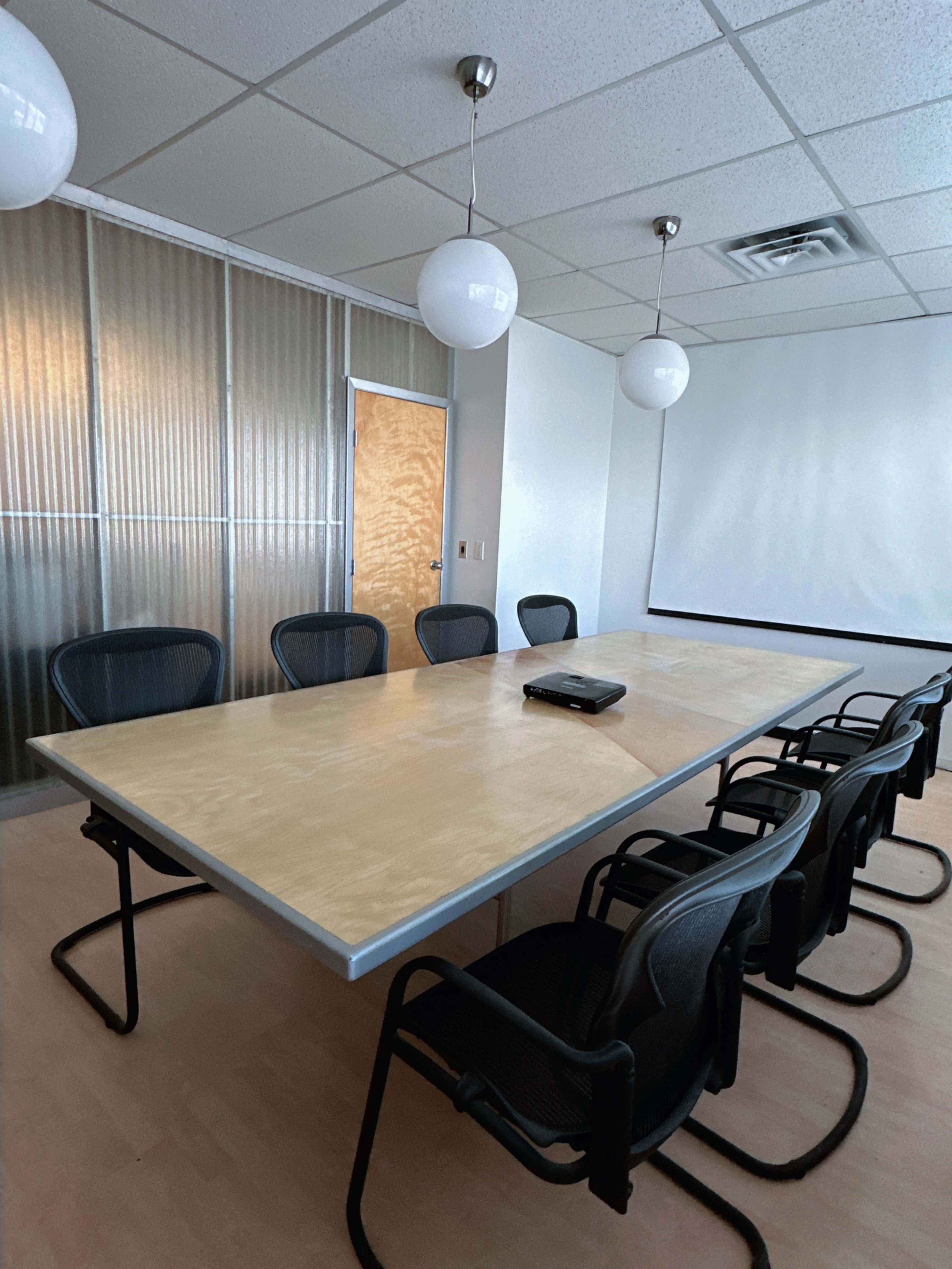 A conference room features a large rectangular table surrounded by black mesh chairs, with a projector screen and glass partition walls in the background.