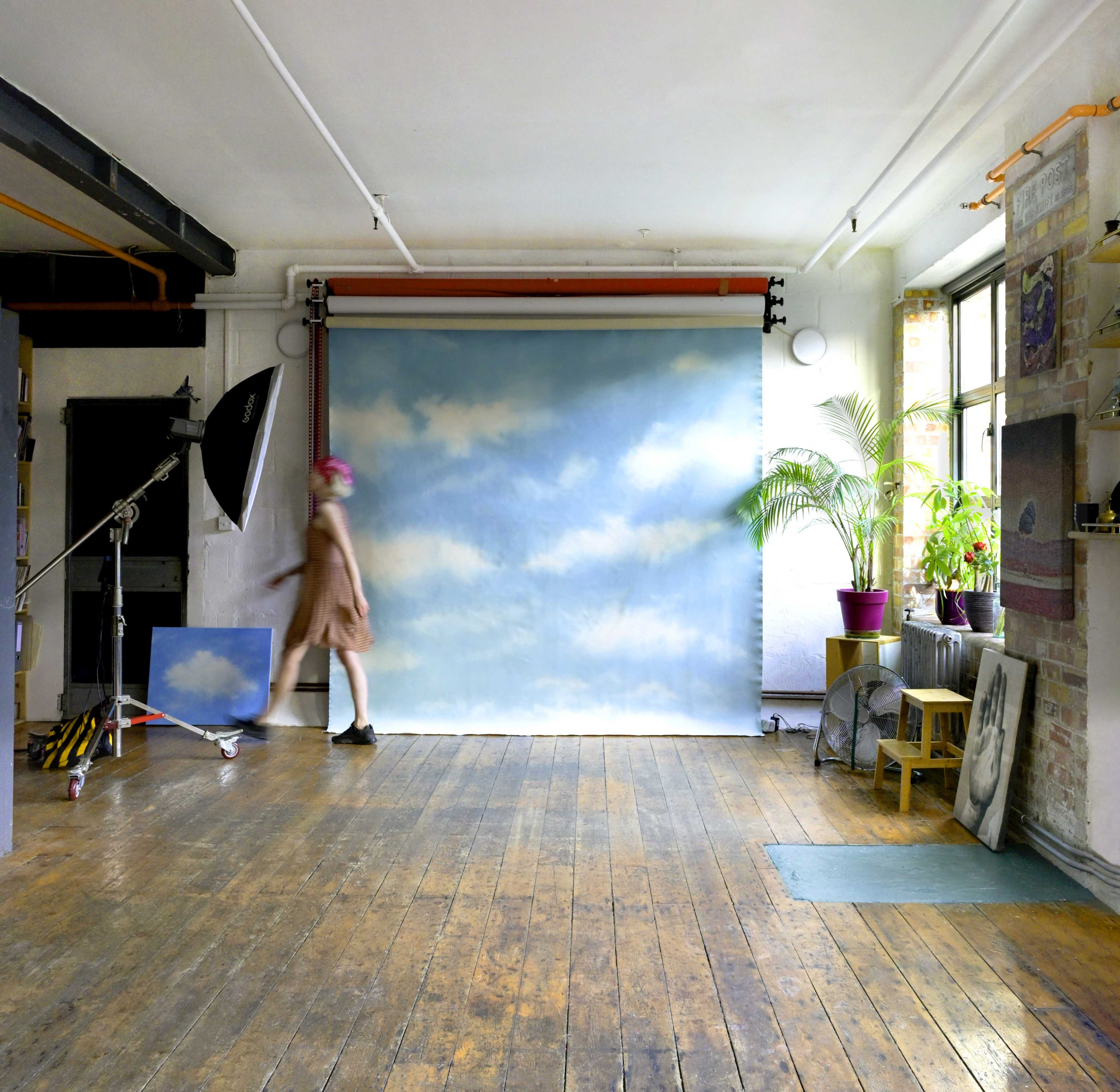 A person walks past a blue sky backdrop in a spacious studio with wooden floors and a potted plant.