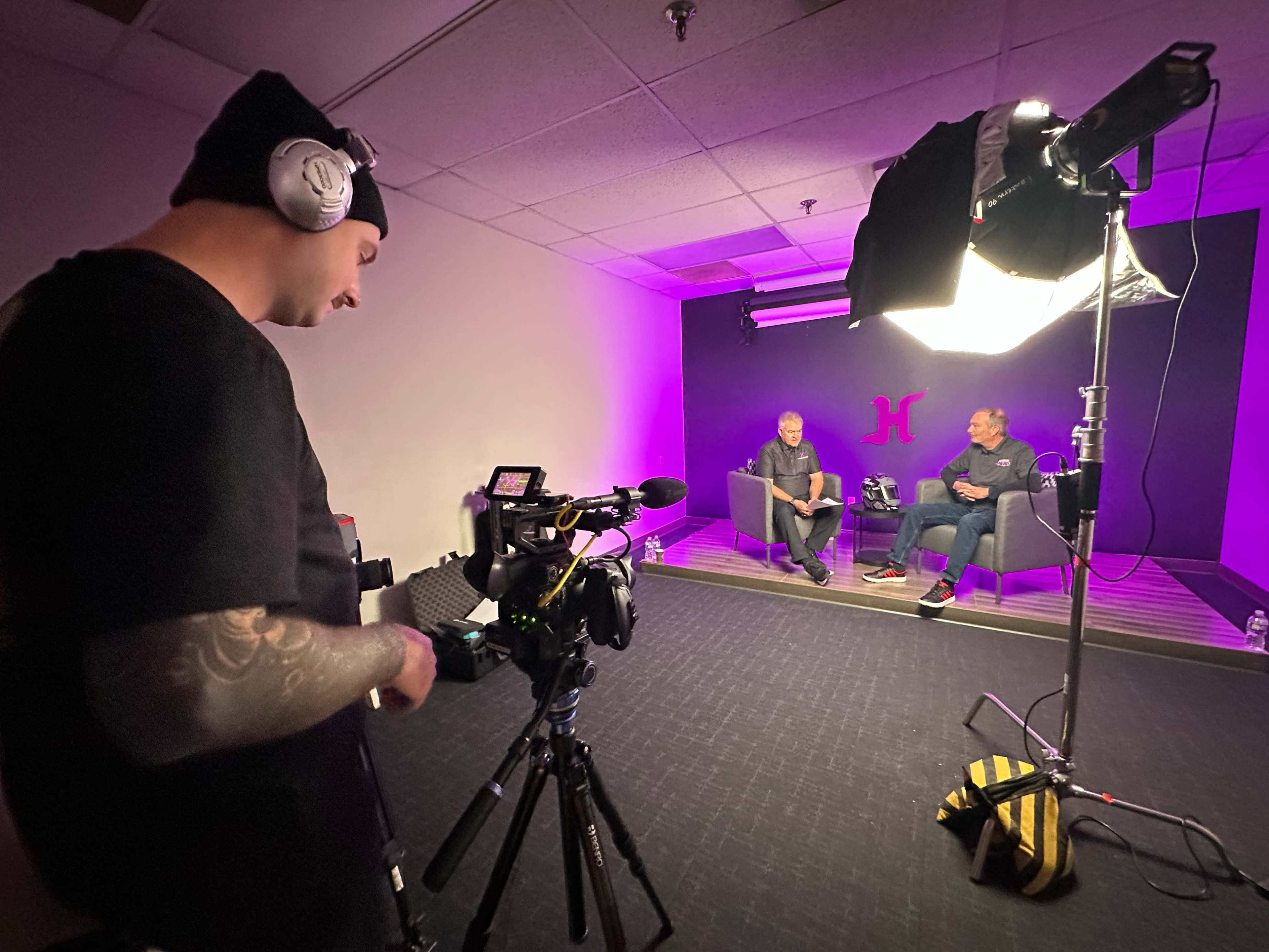 A cameraman operates a video camera while two men sit on chairs in a low-lit studio with purple lighting.