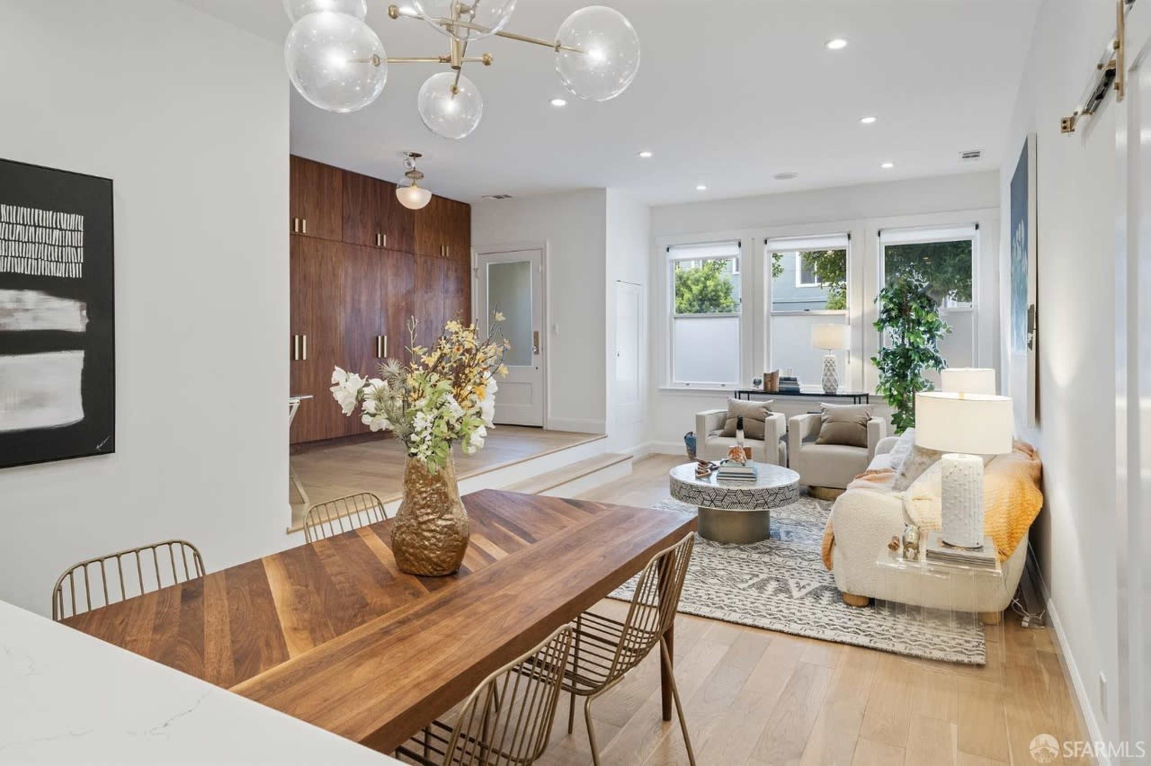 A modern living space featuring a dining area with a wood table, a bright sitting area with a couch and coffee table, and a kitchen with wooden cabinetry.