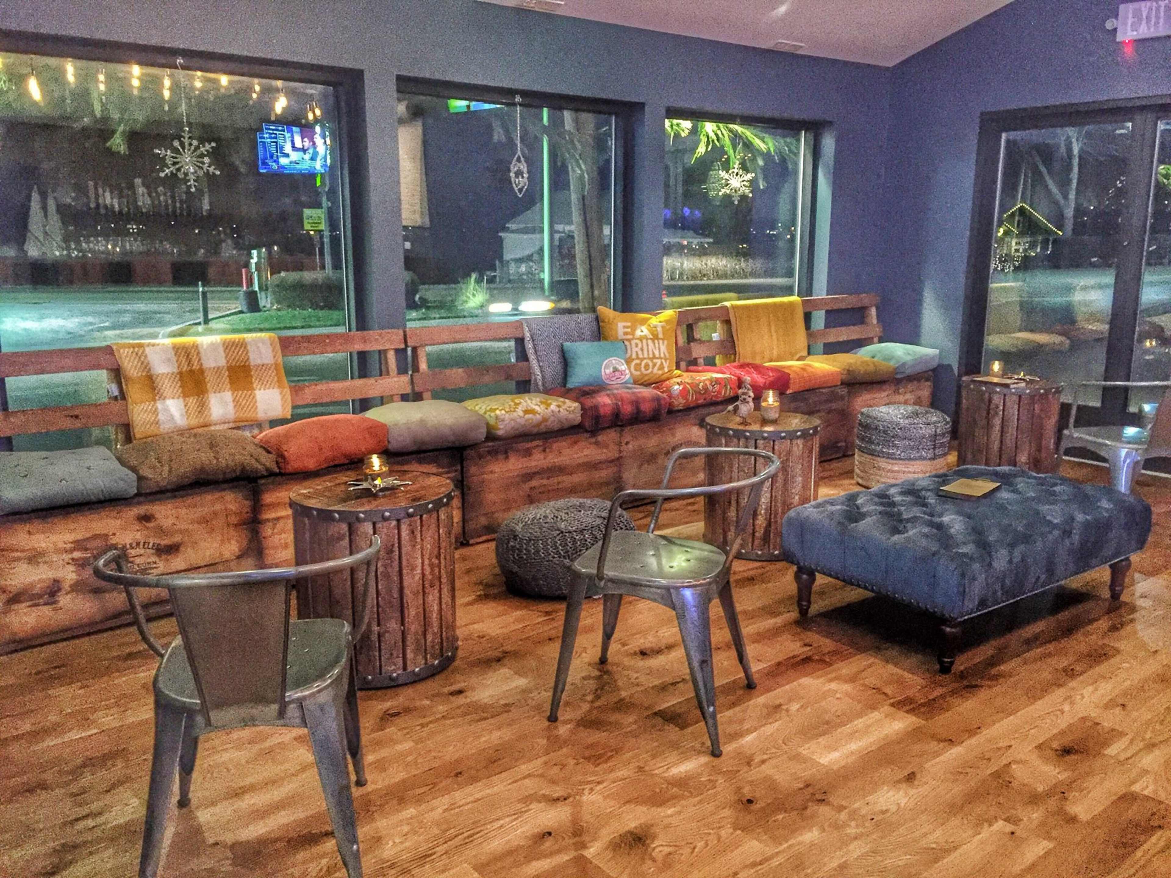 The image shows a cozy seating area with wooden benches lined with colorful cushions, metal chairs, and rustic side tables in a warmly lit room.