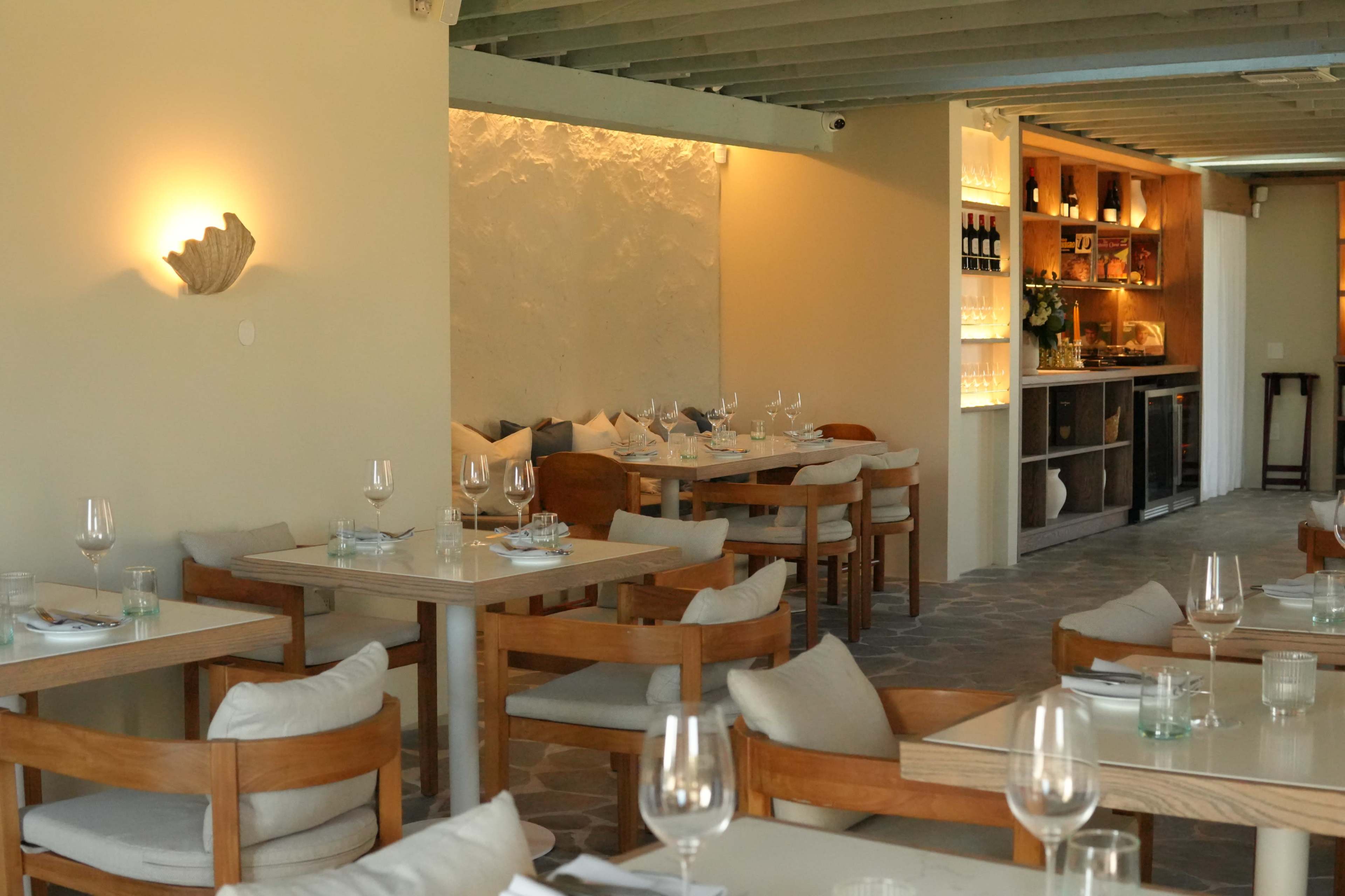 The image shows a modern restaurant with wooden tables set for dining, featuring soft lighting and a minimalist decor.