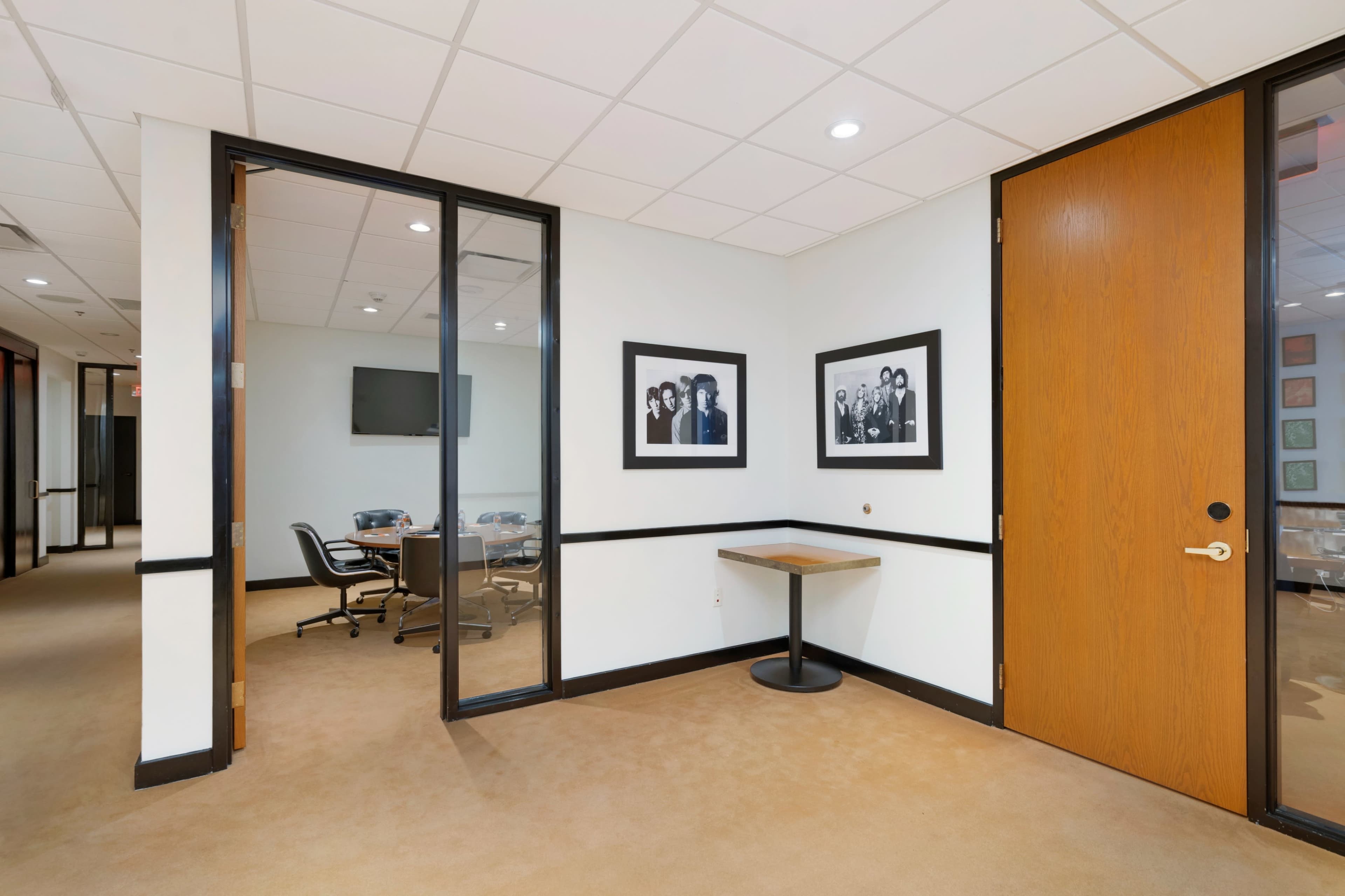 The image shows an office hallway with a glass-enclosed meeting room, a wooden door, and two framed black-and-white photographs on the wall.