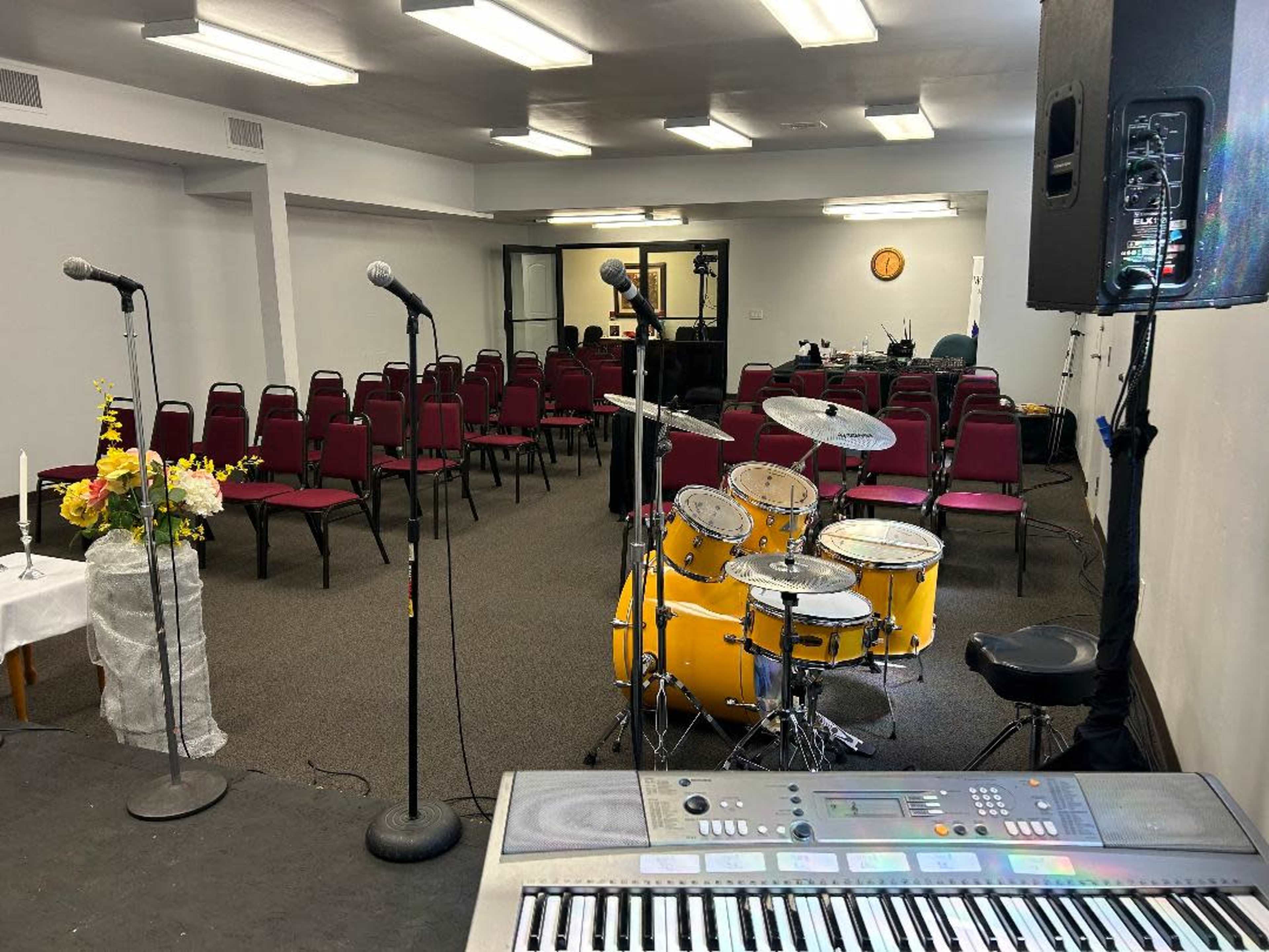 A setup for a performance or meeting, featuring a stage with a keyboard, a drum set, microphones, and rows of empty chairs arranged in a room.