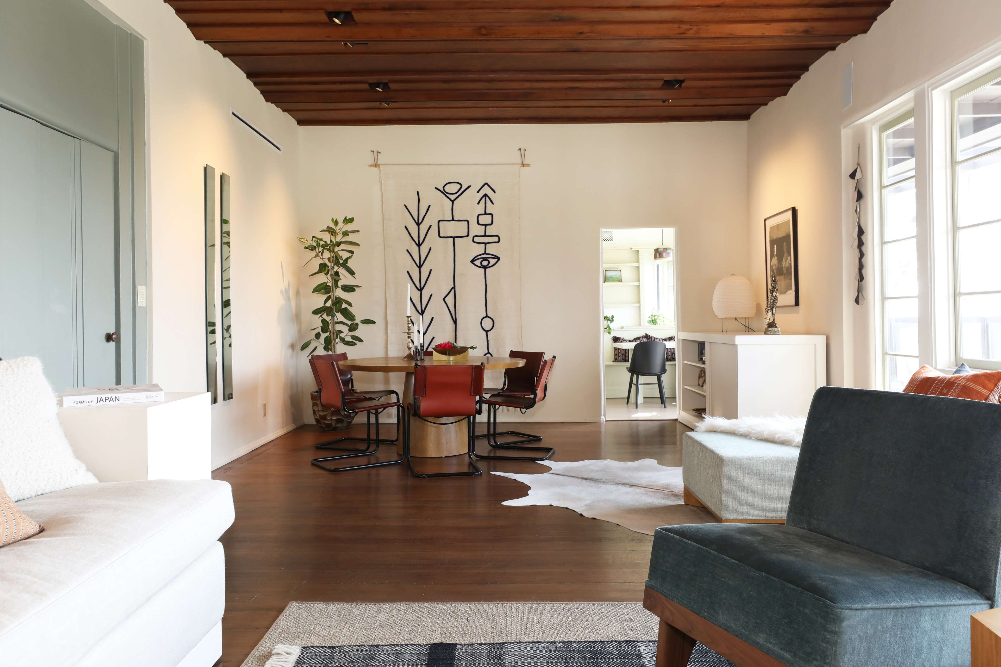 A living room with wooden beams, a cozy couch, and a dining area featuring a patterned wall hanging and a dining table surrounded by chairs.