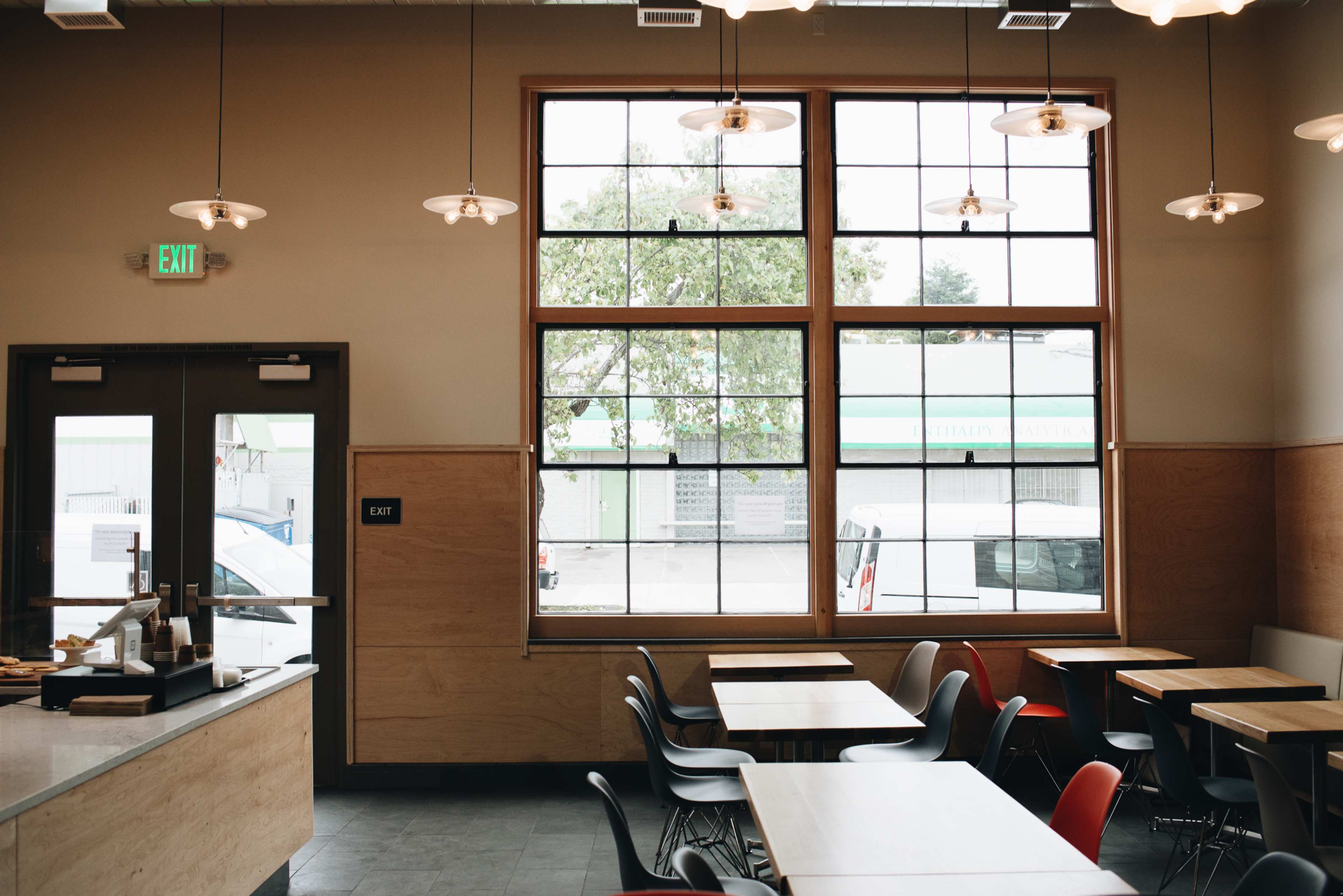 A restaurant interior features wooden tables and chairs with large windows allowing natural light to enter.