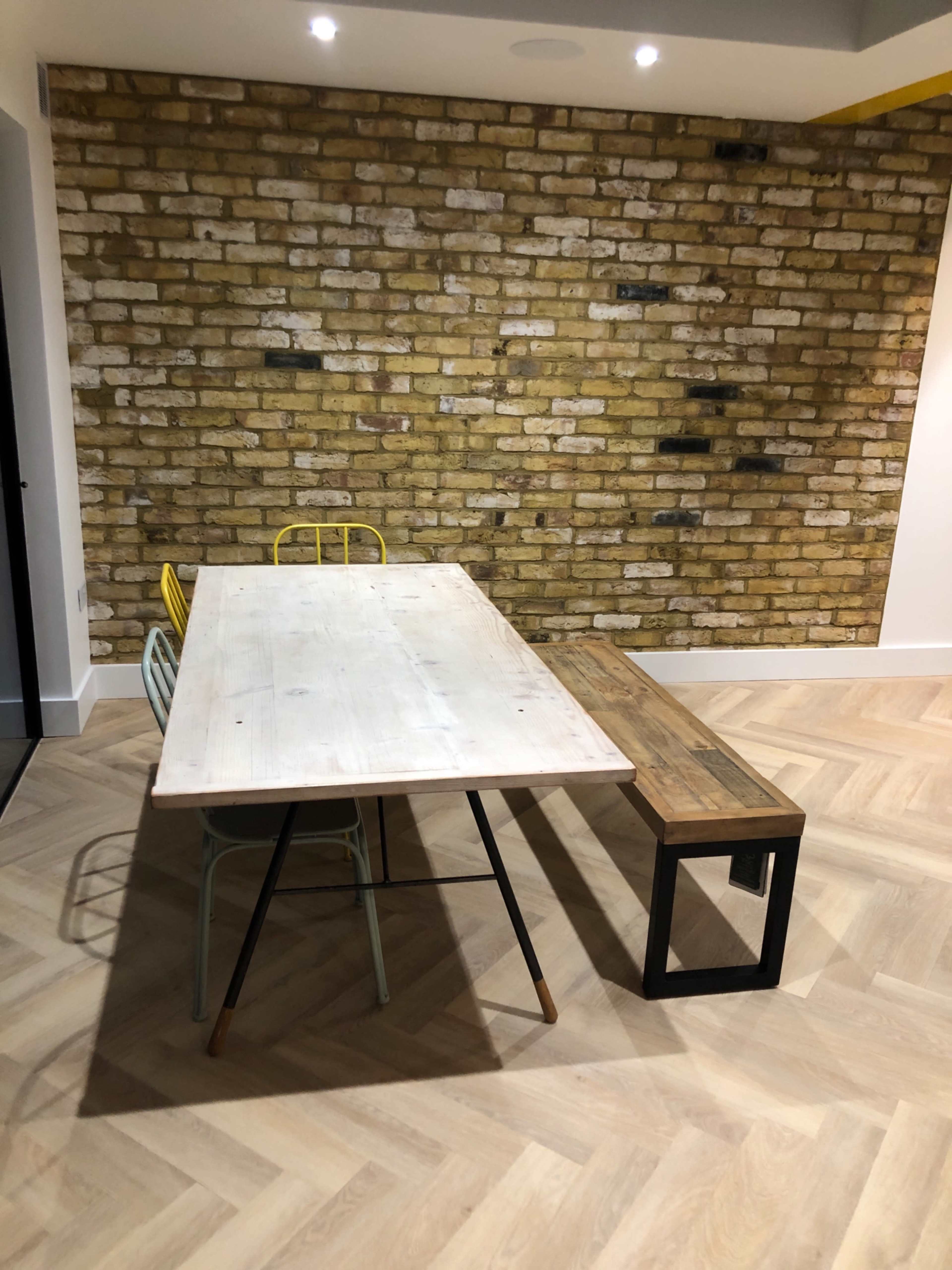A wooden dining table is positioned in front of a brick wall, accompanied by a bench and several metal chairs.
