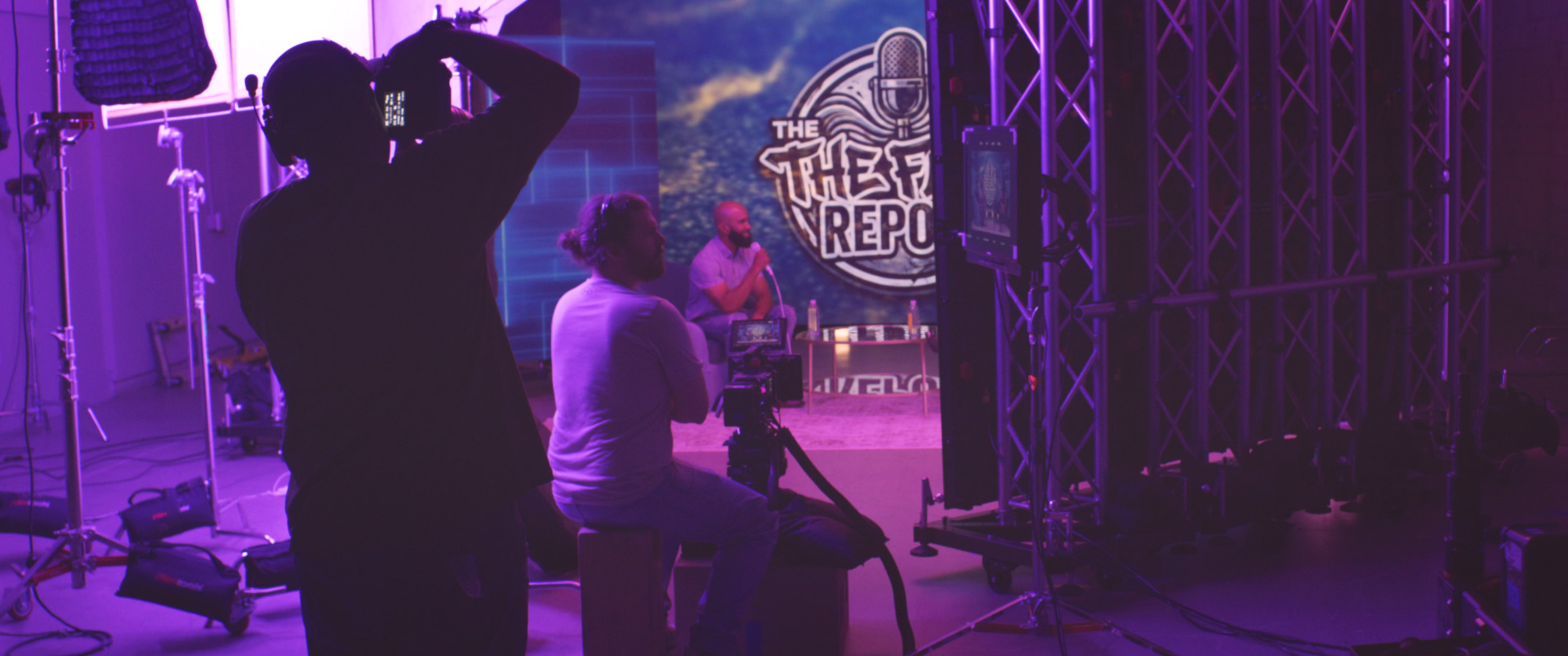 A camera operator captures a scene with a host seated in front of a backdrop featuring "The F Report" during a studio production.