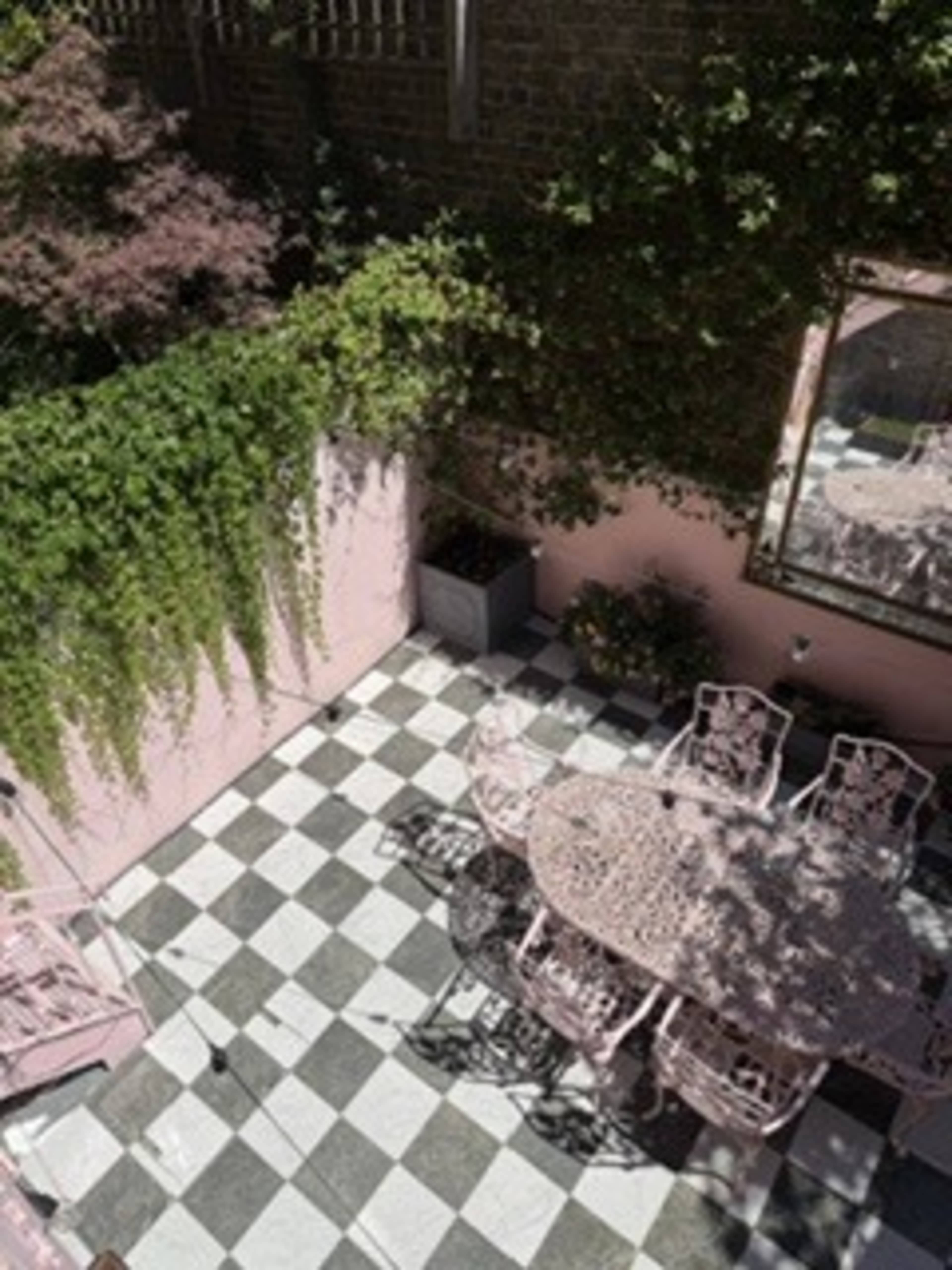 The image shows a patio area with a round, pink wrought-iron table surrounded by matching chairs, set on a black and white checkered floor, with greenery and a mirror on the walls.