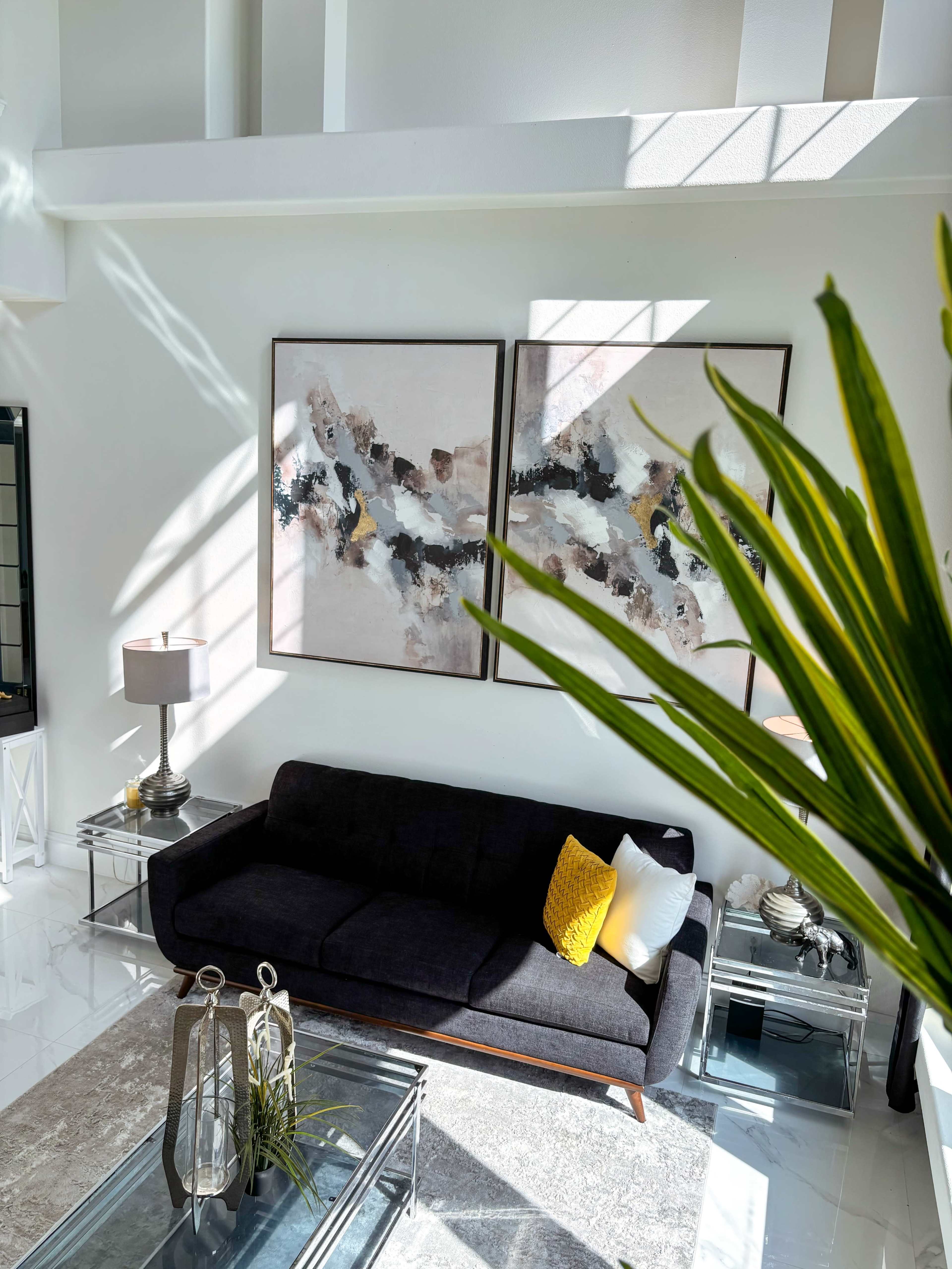 A modern living room features a dark sofa, two large abstract paintings on the wall, and a glass coffee table surrounded by decorative items and a potted plant.
