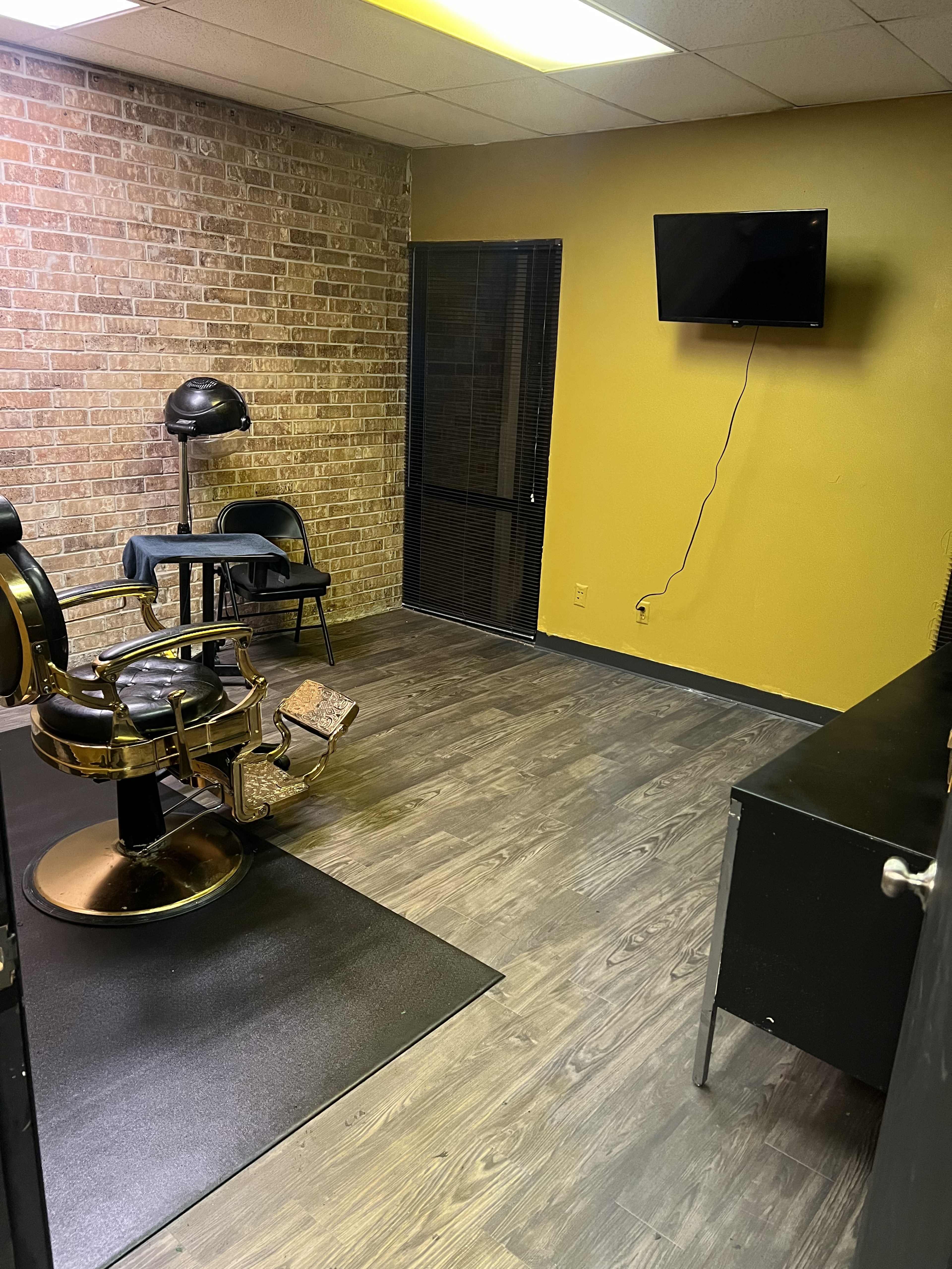 A room with a barber chair, a small desk, and a wall-mounted television, featuring brick walls and a yellow paint accent.