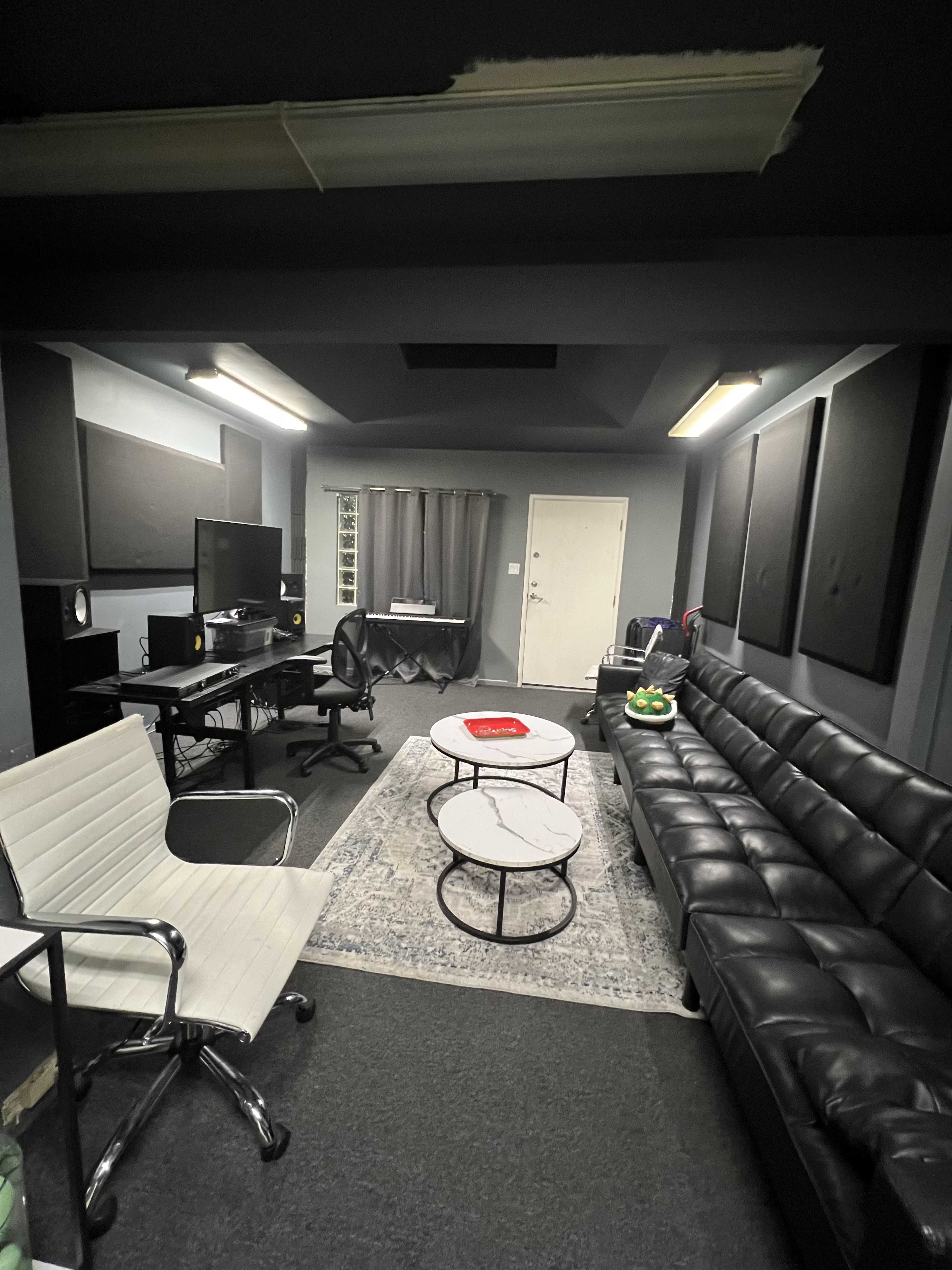 A small, modern office or studio space featuring a black leather couch, a couple of round tables, a desk with a computer, and soundproofing panels on the walls.