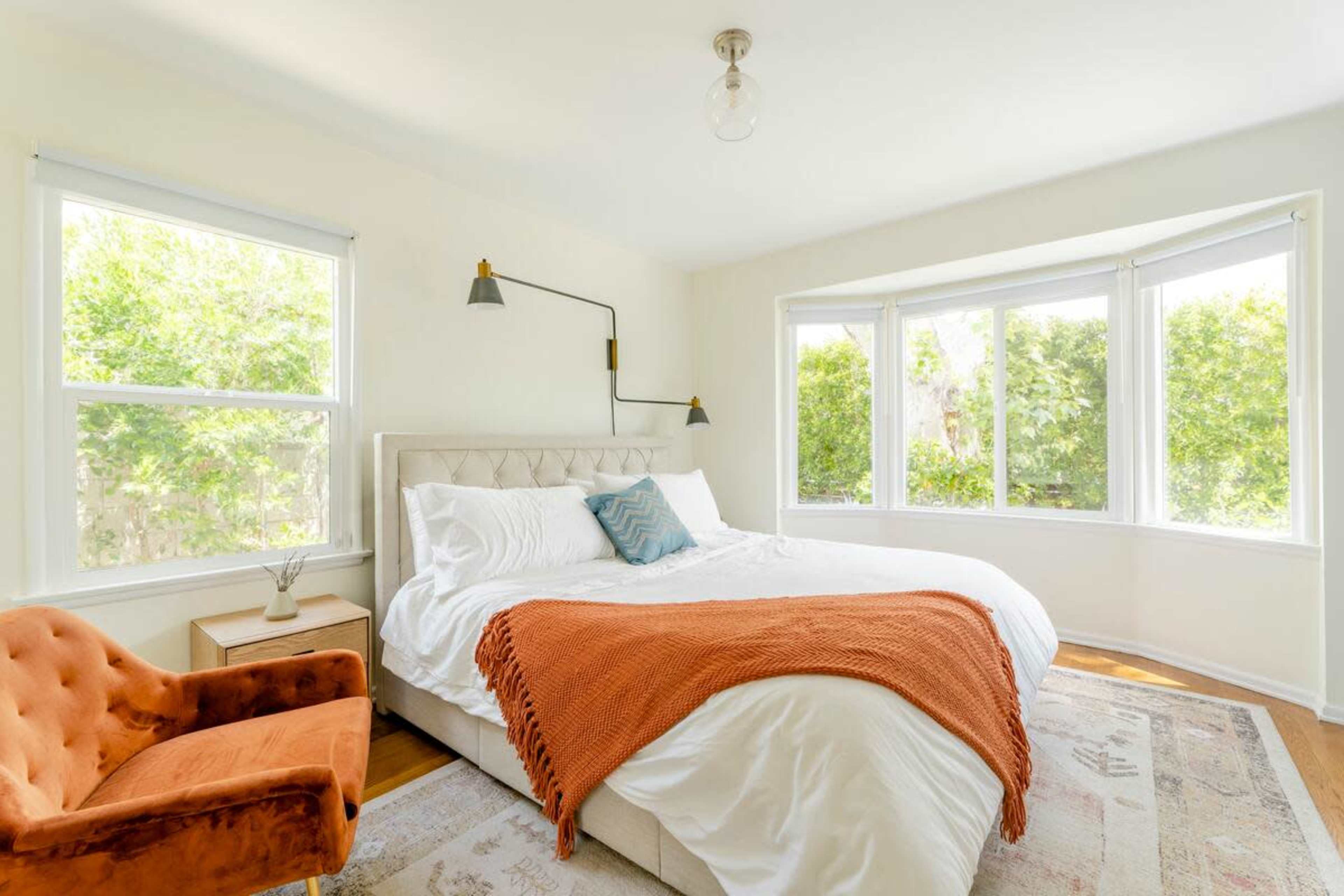 The bedroom features a large bed with a white comforter, an orange throw blanket, a cozy armchair, and large windows letting in natural light.