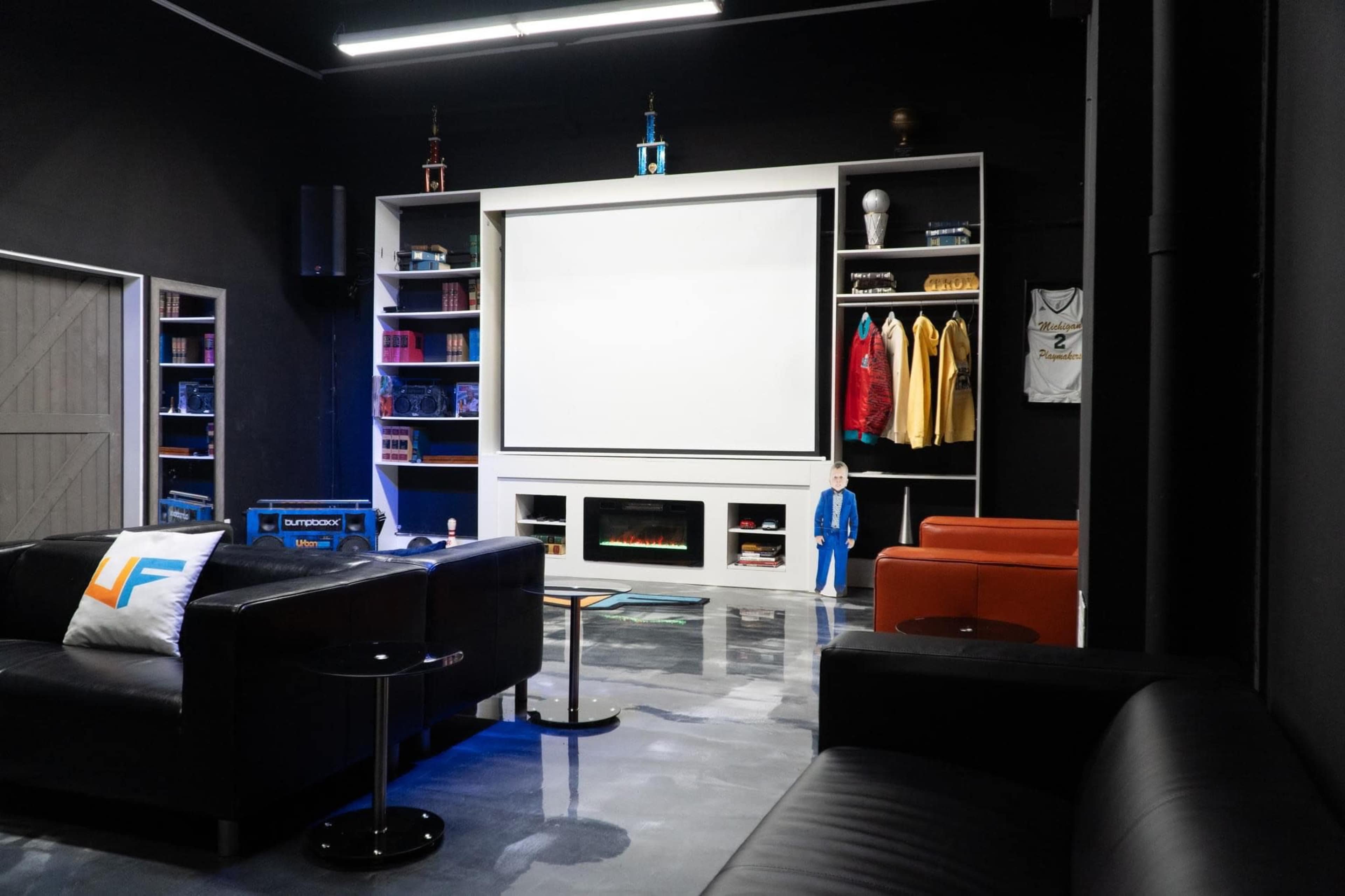 The image shows a modern entertainment room with a large projection screen, a sleek black couch, and shelves displaying various items, including sports jerseys and trophies.