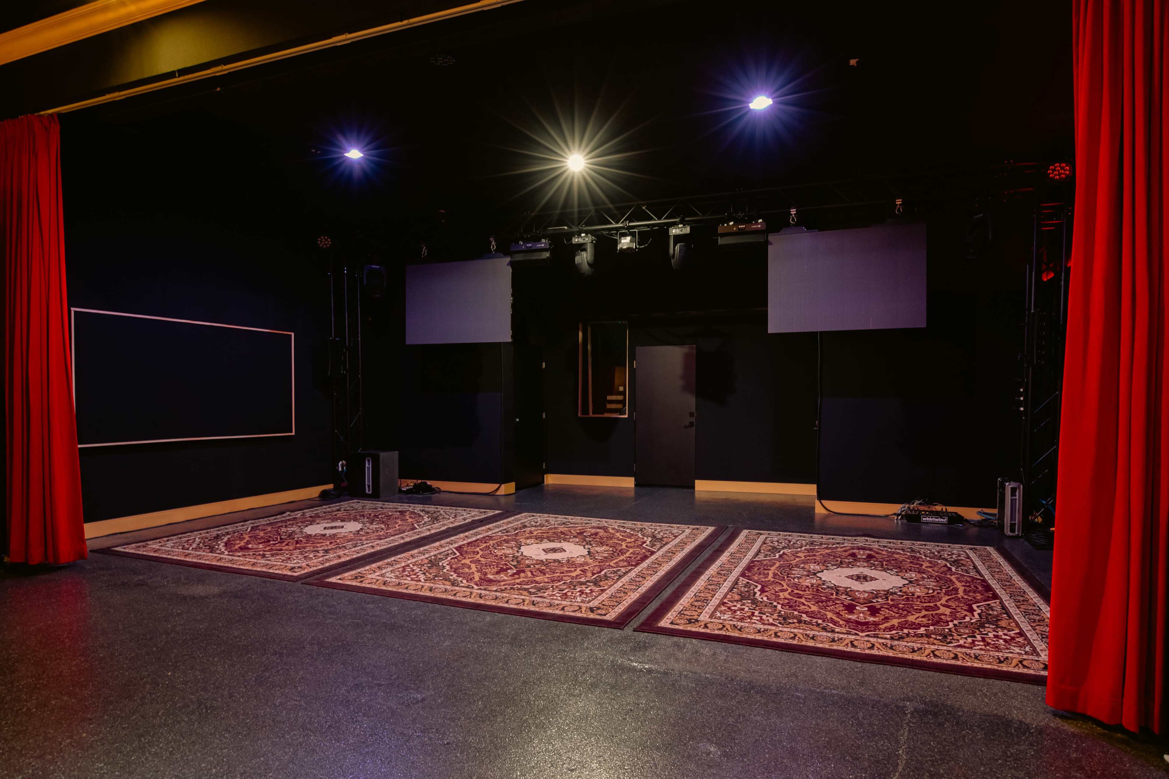 The image shows an empty stage with three large rugs, spotlights overhead, and red curtains framing the sides.