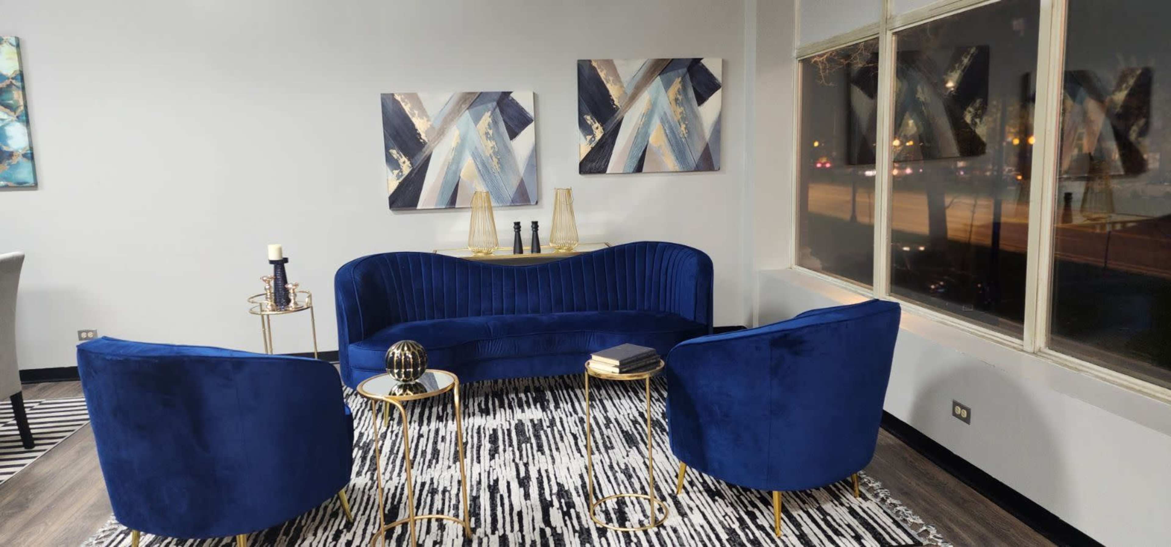 A modern lounge area features a blue velvet sofa and two matching armchairs arranged around a circular gold side table on a patterned rug.
