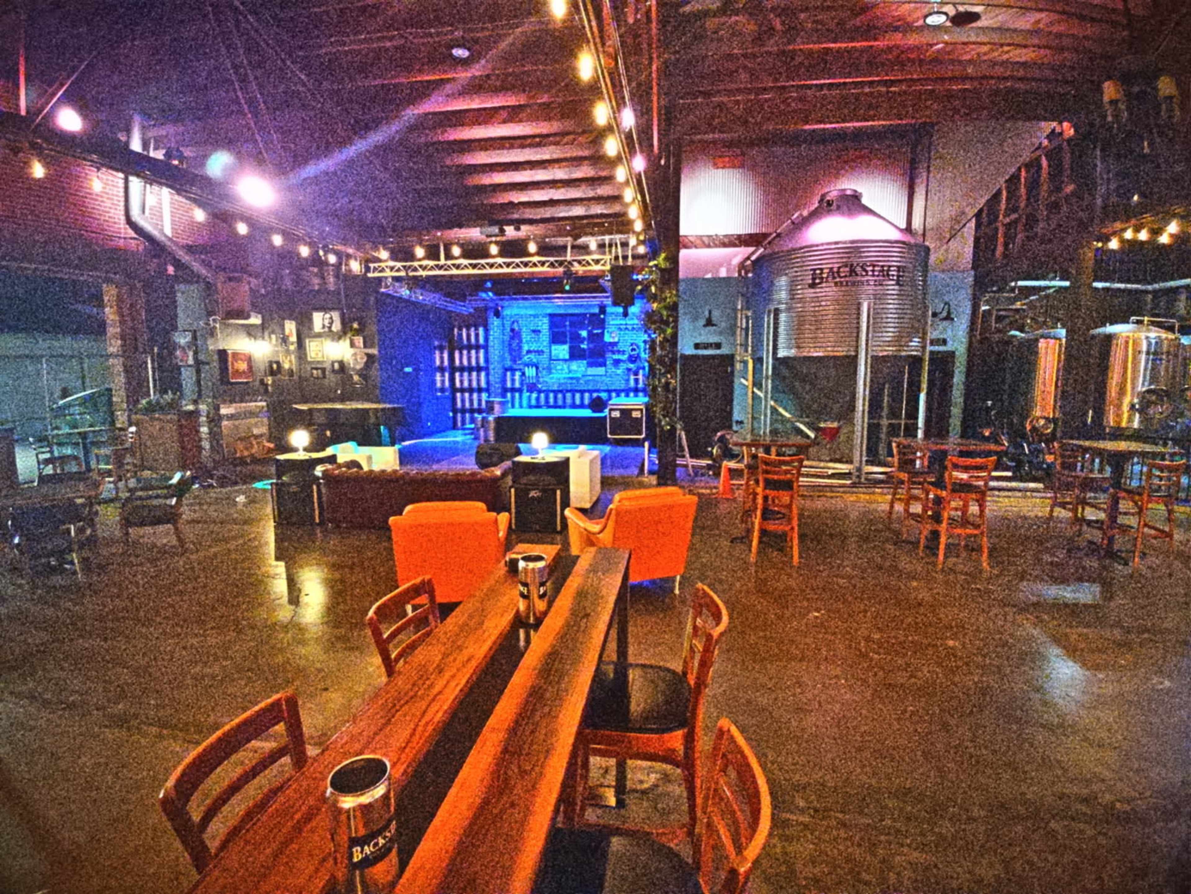 The image shows a spacious, industrial-style bar or brewery with wooden tables, chairs, and large brewing tanks in the background.