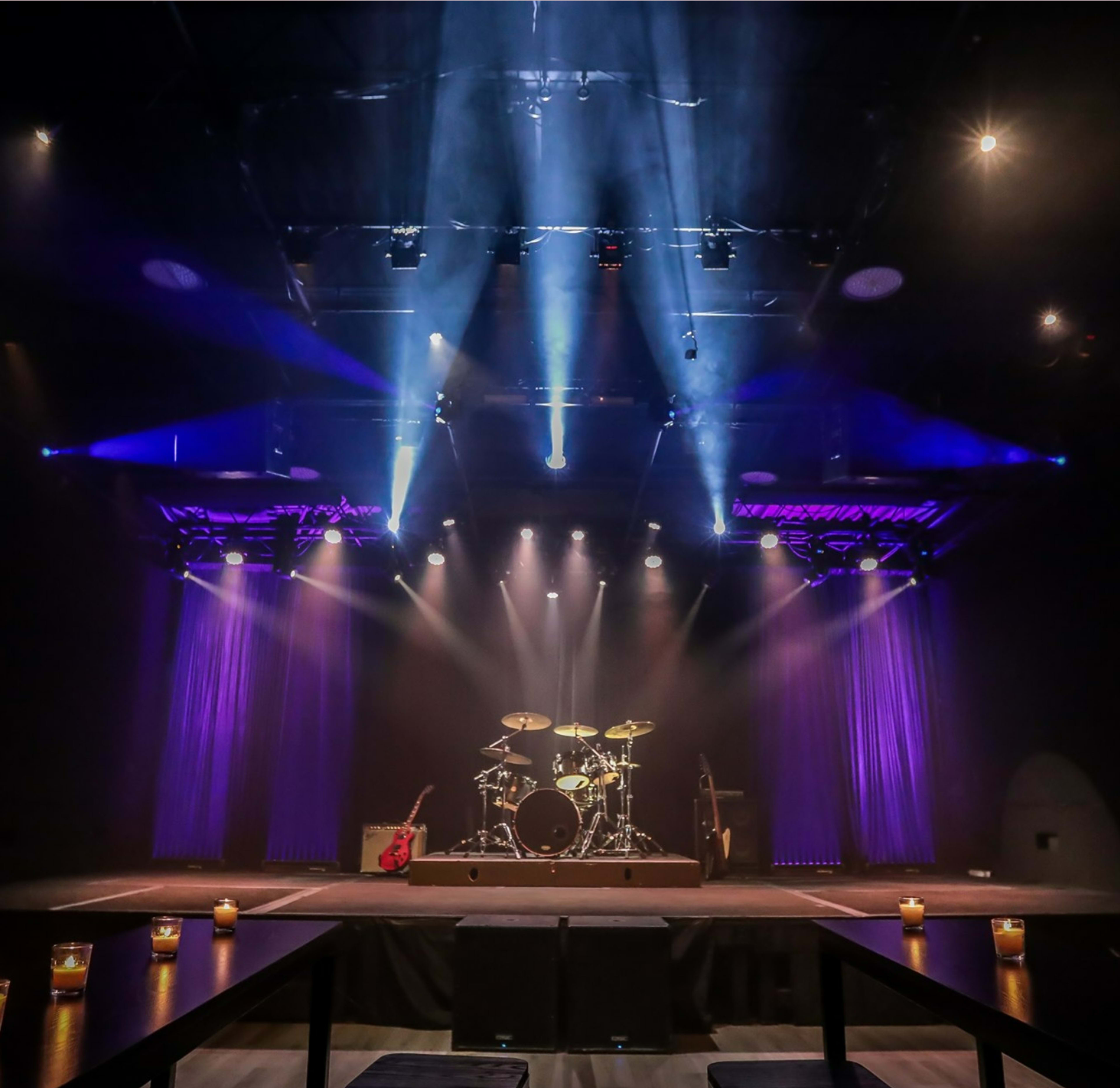 A stage is set with a drum kit and electric guitar, illuminated by blue and purple stage lights, surrounded by candlelit tables.