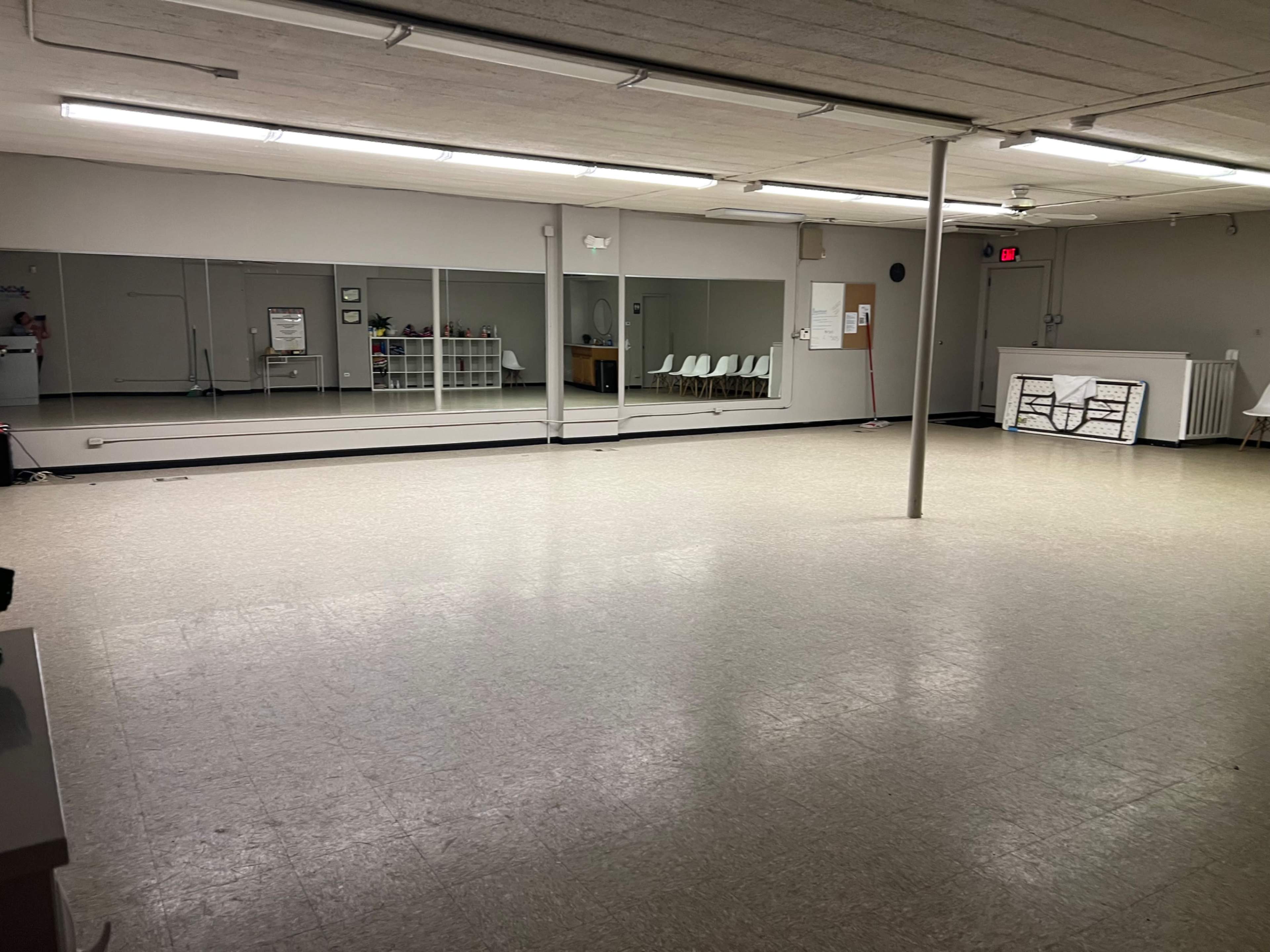 The image shows a spacious, empty interior room with mirrored walls and a polished floor, featuring a few chairs and storage furniture.