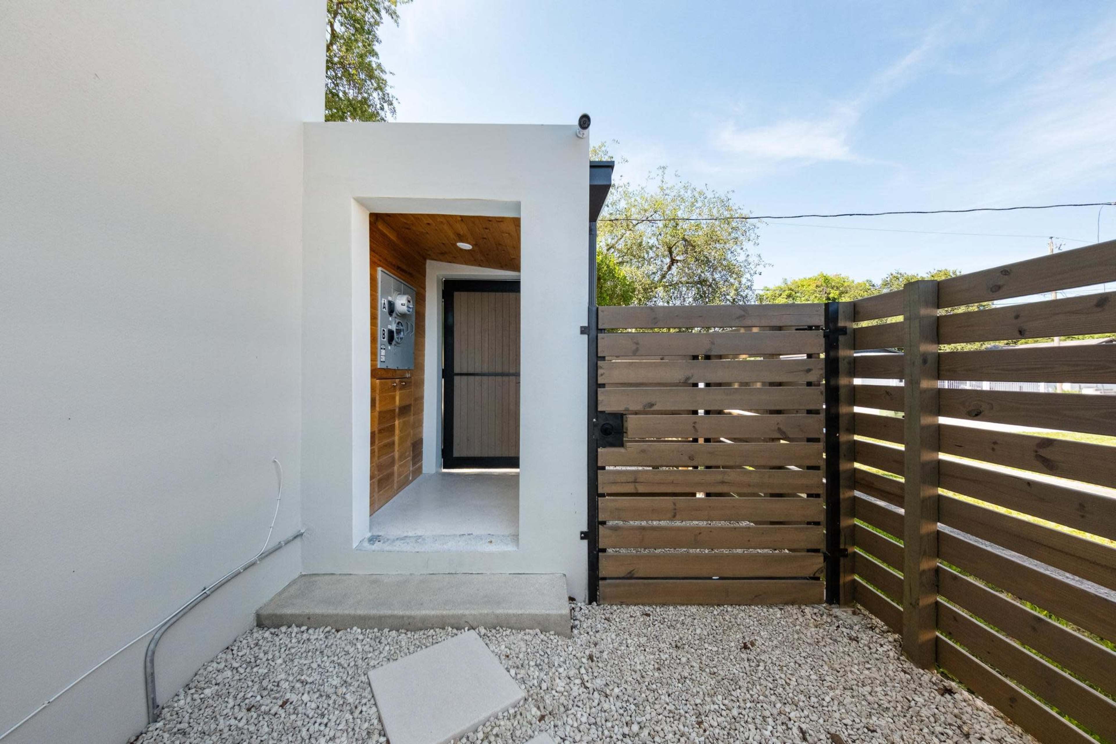 The image shows a small entrance area with a wooden door, surrounded by a fenced yard featuring gravel and a paved entrance.