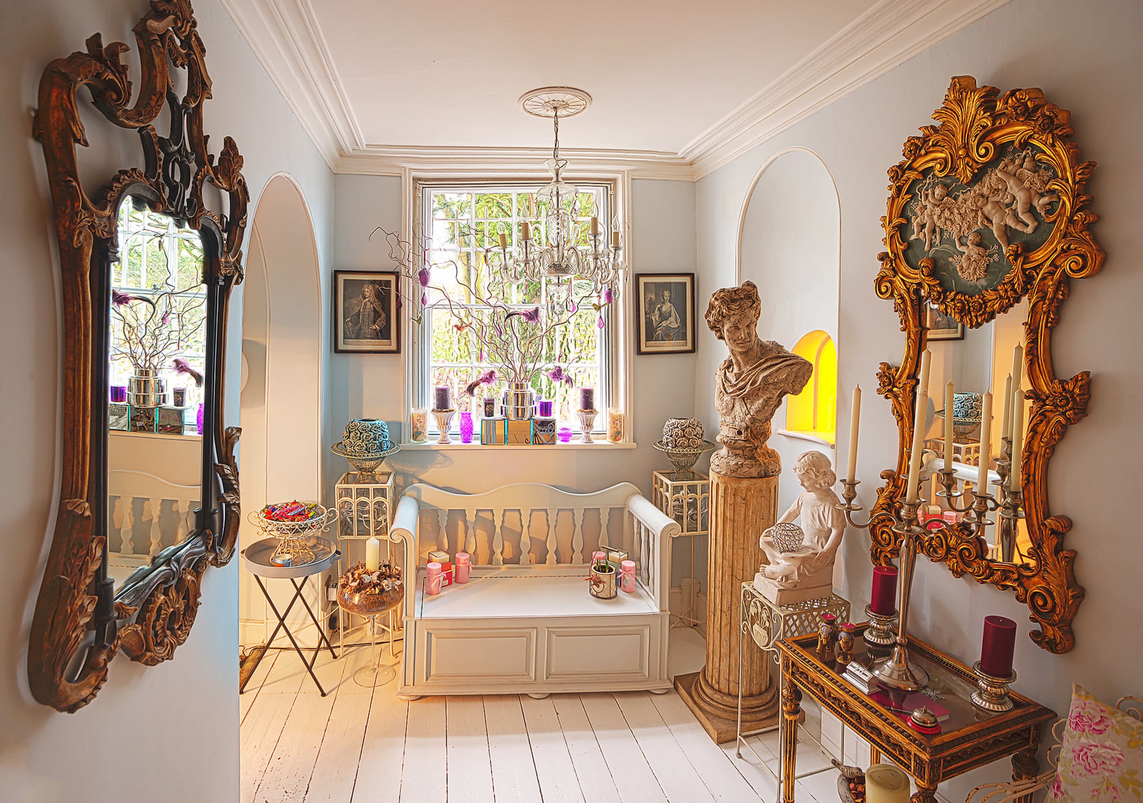 The image shows a bright hallway adorned with ornate mirrors, sculptures, and decorative furnishings.