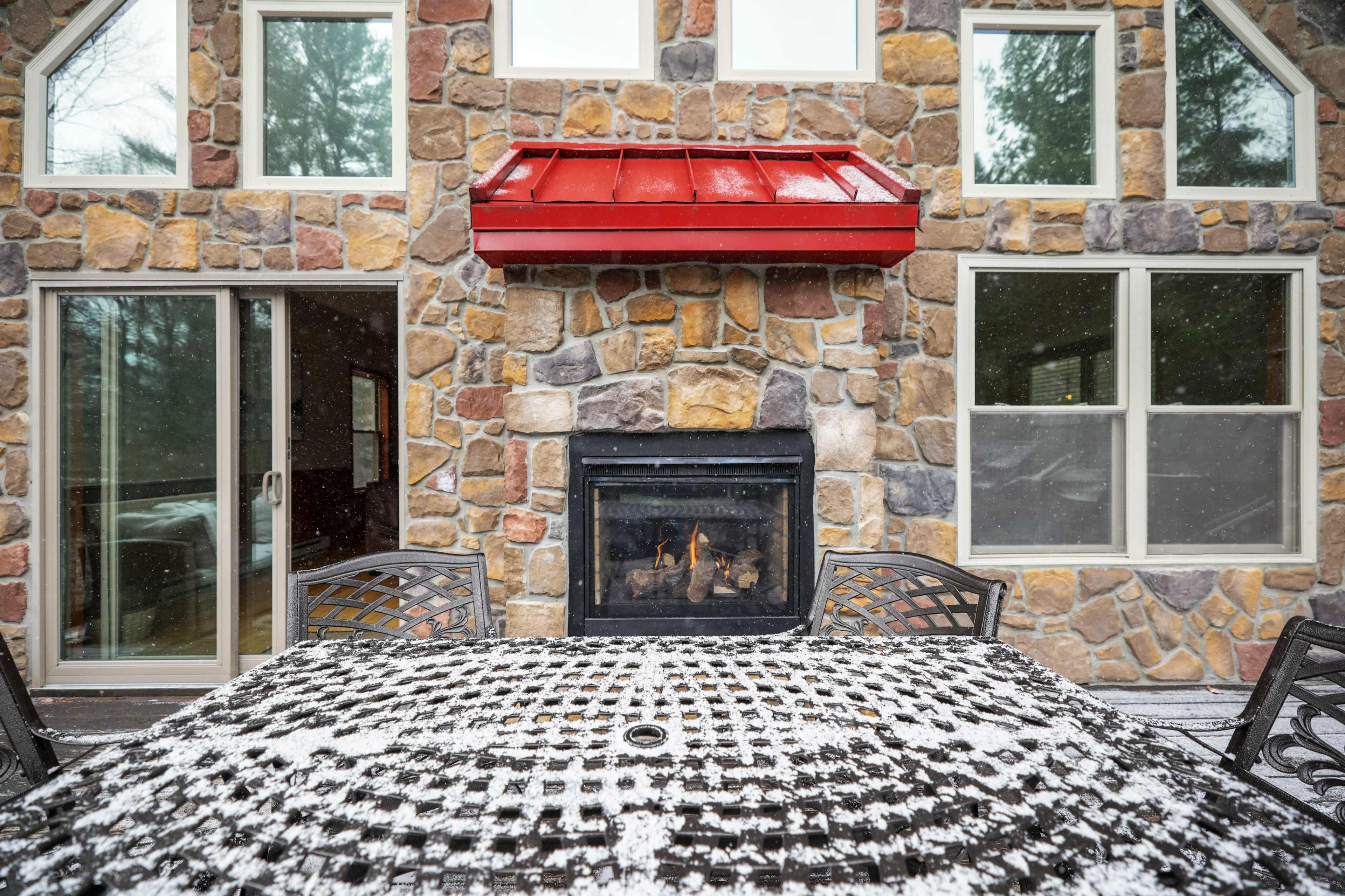 A stone exterior features a red metal roof, with a fireplace and a table covered in snow in an outdoor seating area.