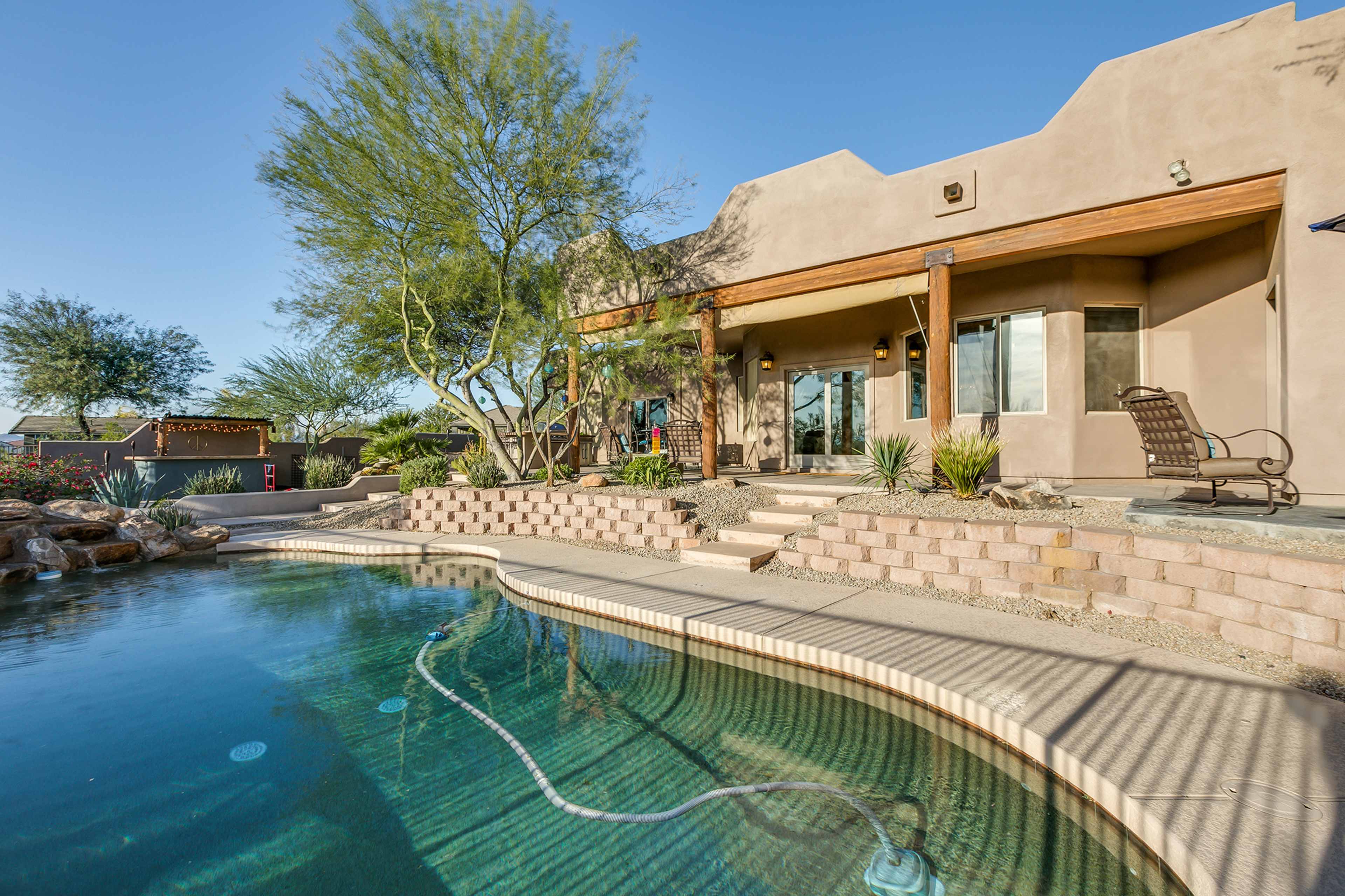 A home with a stucco exterior is situated beside a swimming pool, surrounded by desert landscaping and a clear blue sky.