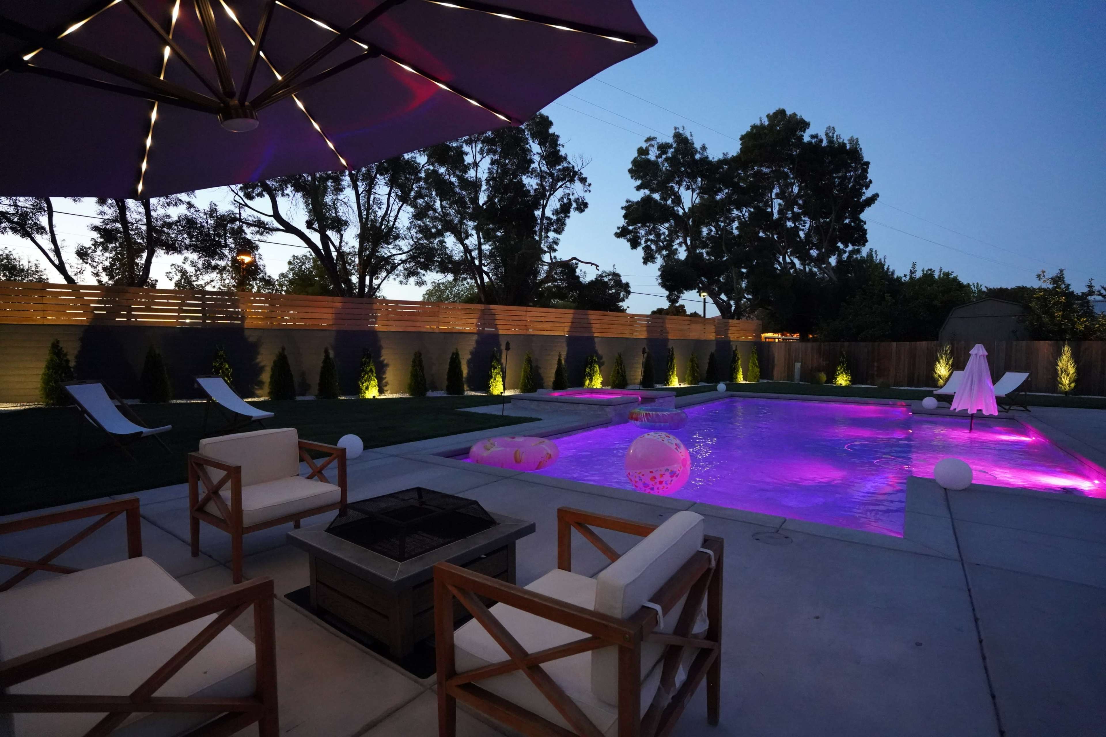 A backyard pool area features glowing purple lights, lounge chairs, and an umbrella beside a fire pit.