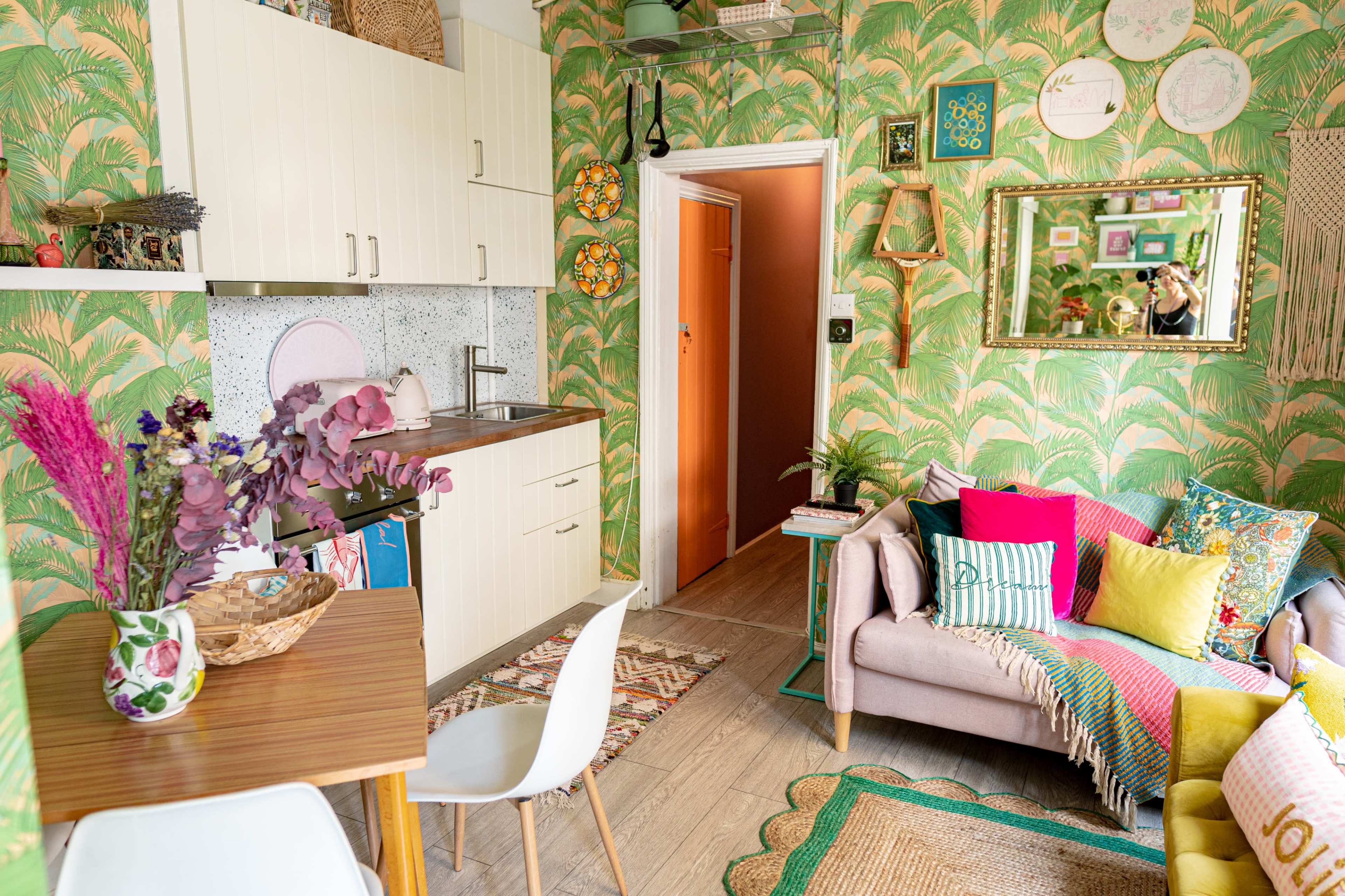 A small, brightly decorated kitchen-living area features tropical-patterned wallpaper, a compact dining table, and a couch adorned with colorful cushions.