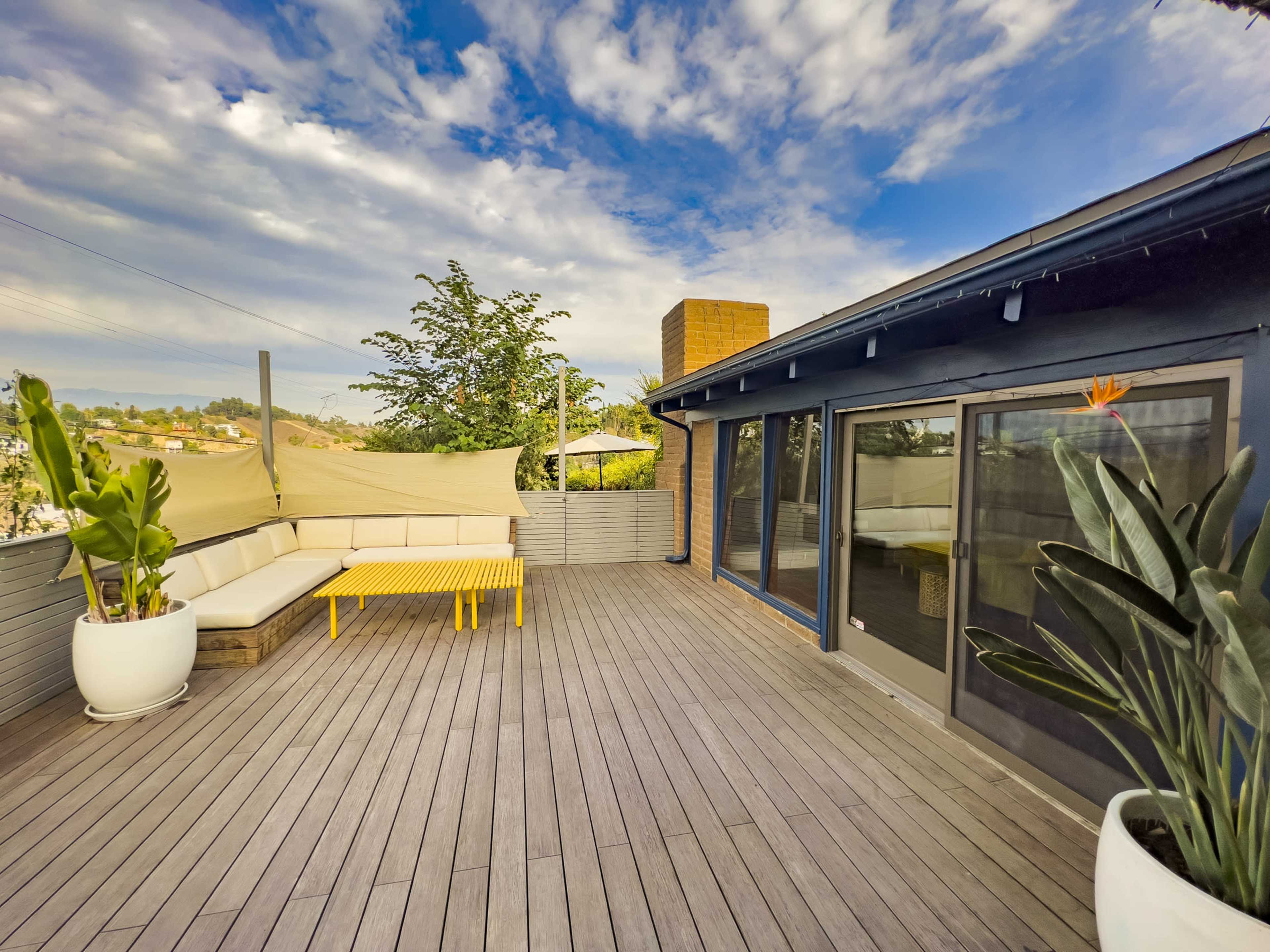 A spacious wooden deck with a yellow table and seating area, surrounded by potted plants under a partly cloudy sky.