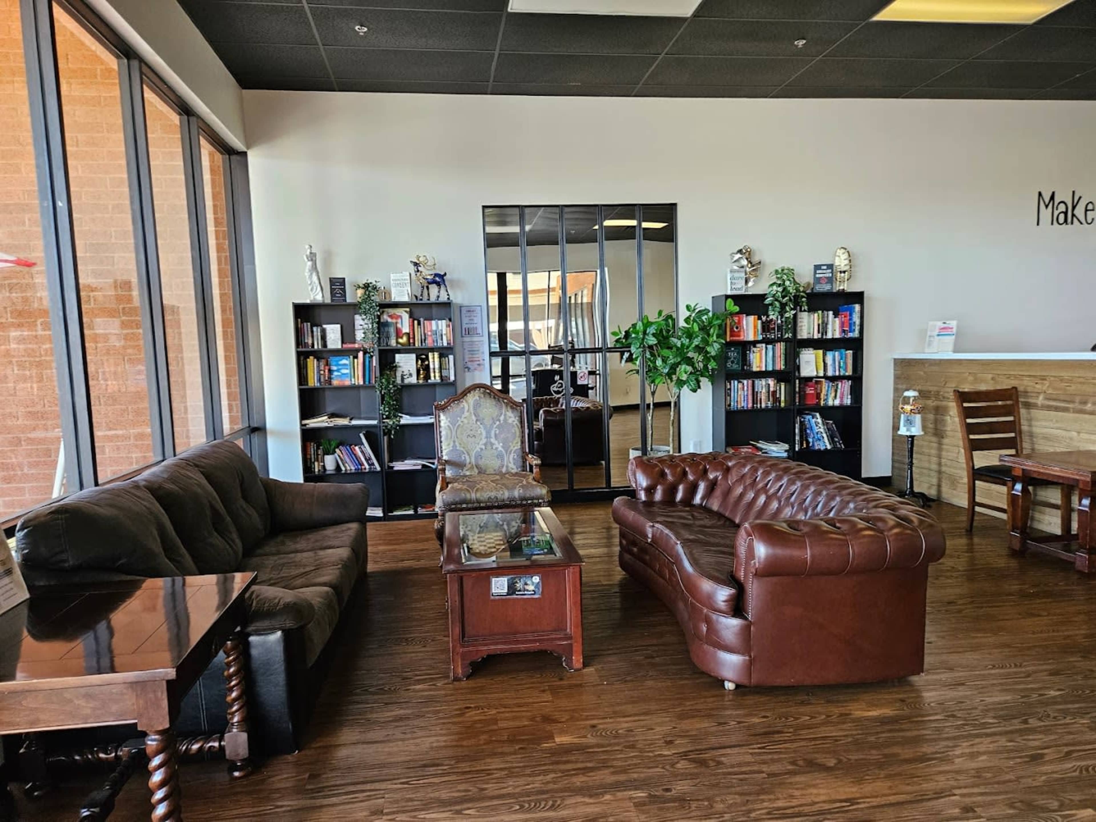 A cozy reading area featuring two sofas, a wooden coffee table, bookshelves filled with books, and large windows allowing natural light.