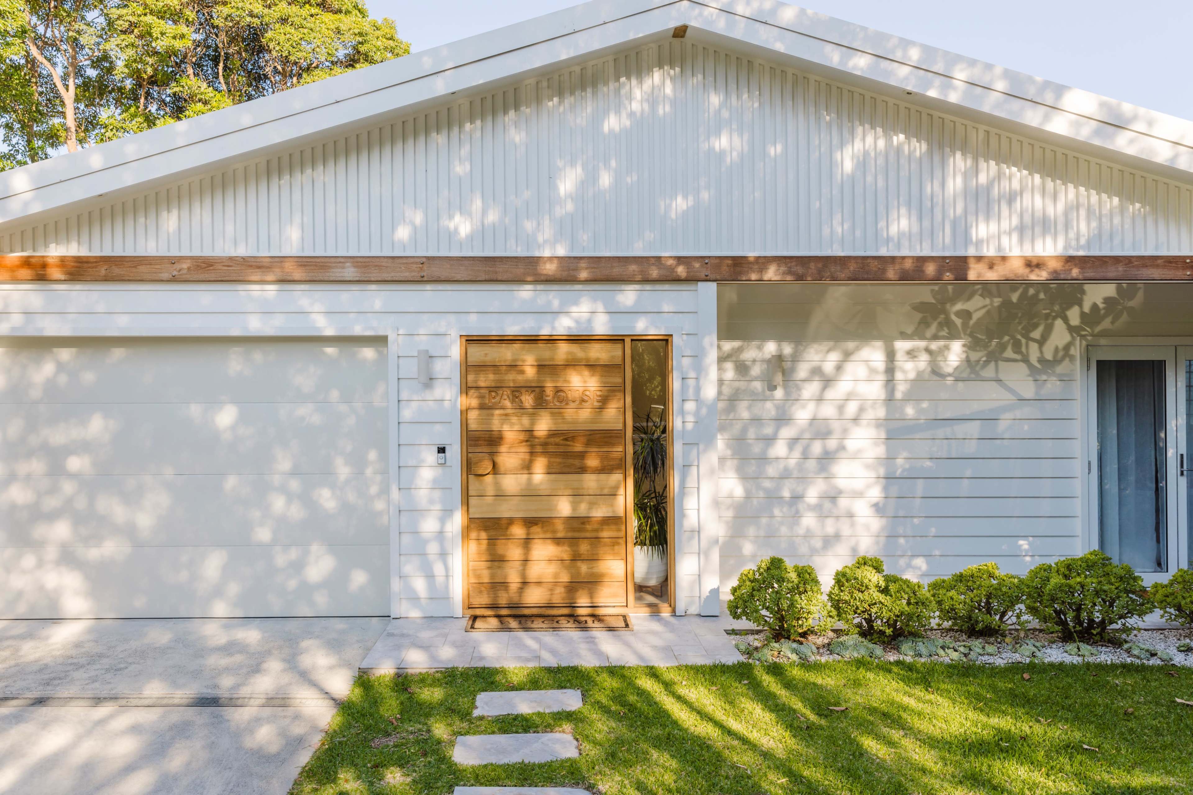 The image shows a modern white house with a wooden front door and a neatly landscaped yard featuring low shrubs and a pathway.