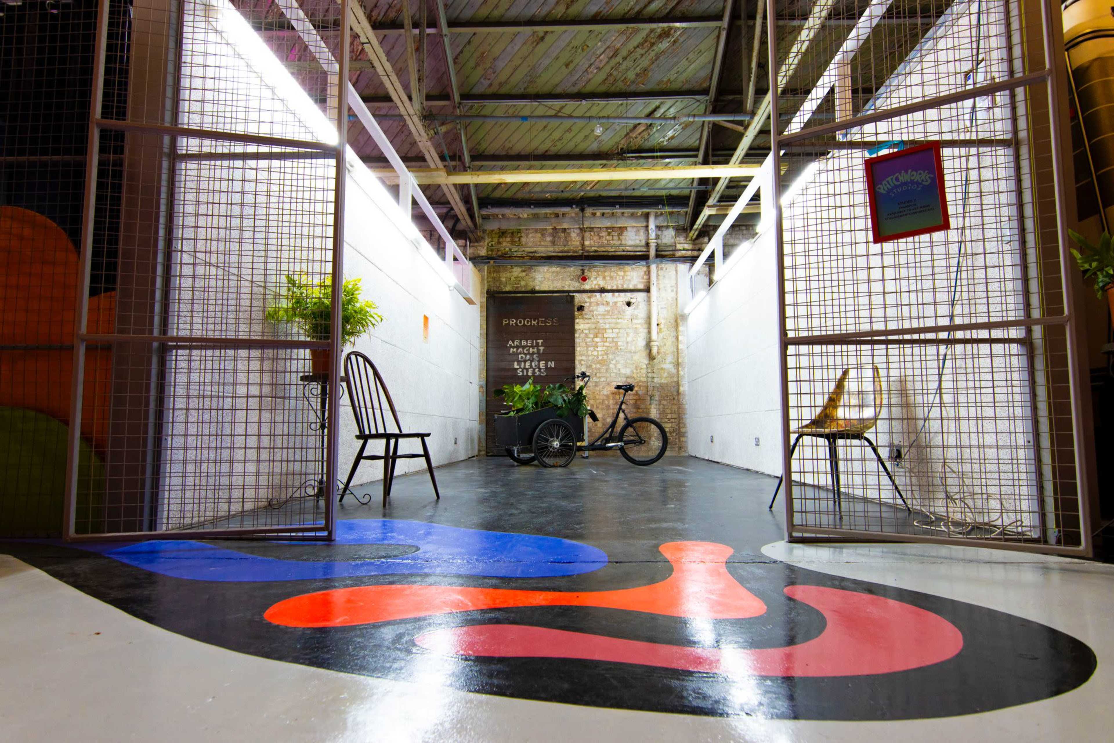 The image shows a narrow hallway with metal gates, a bicycle, and a colorful painted floor, leading to a wall with greenery and artwork.