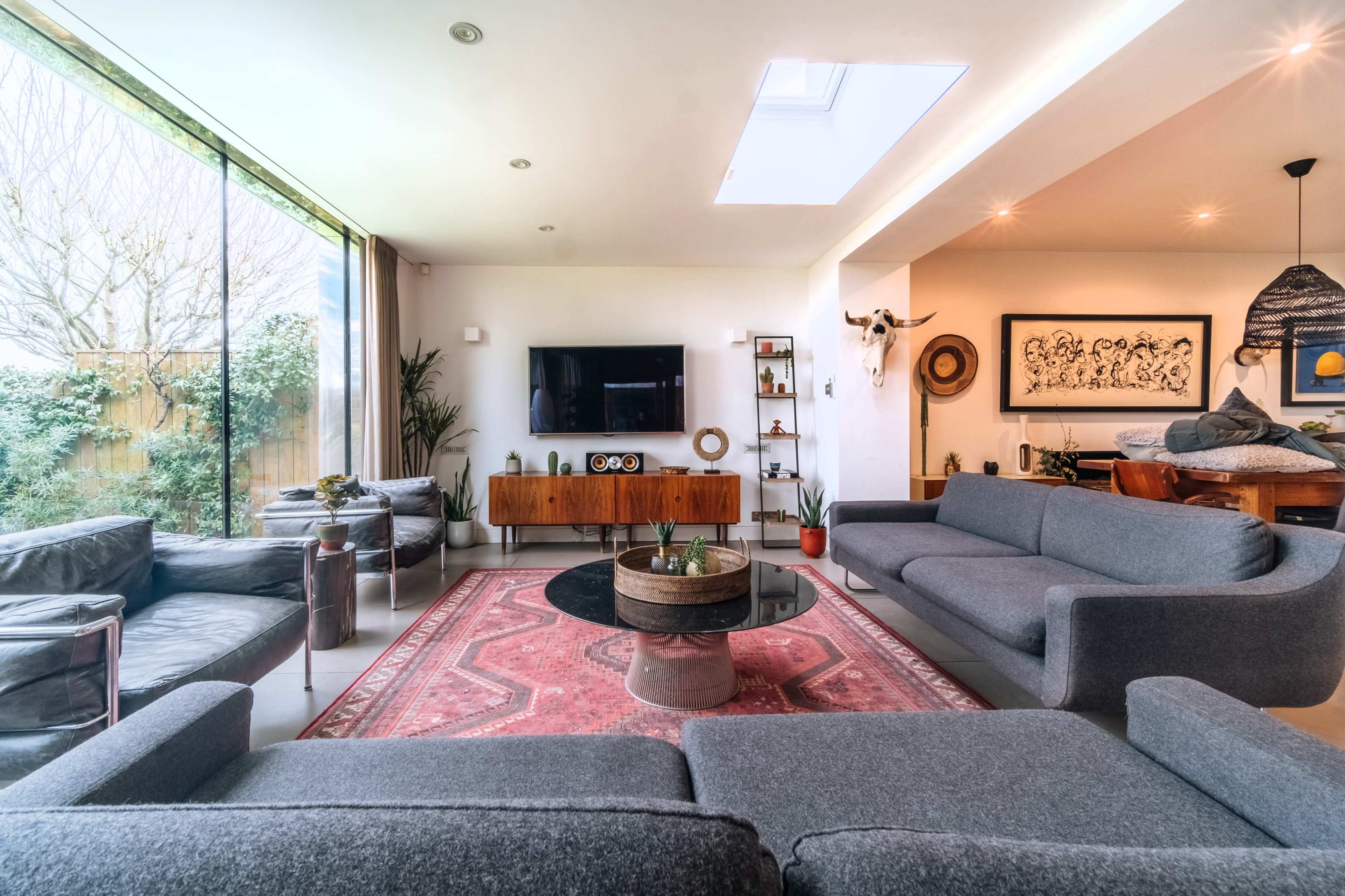 The living room features a large gray sectional sofa, a round black coffee table on a patterned rug, and a wall-mounted television with a window that overlooks a garden.