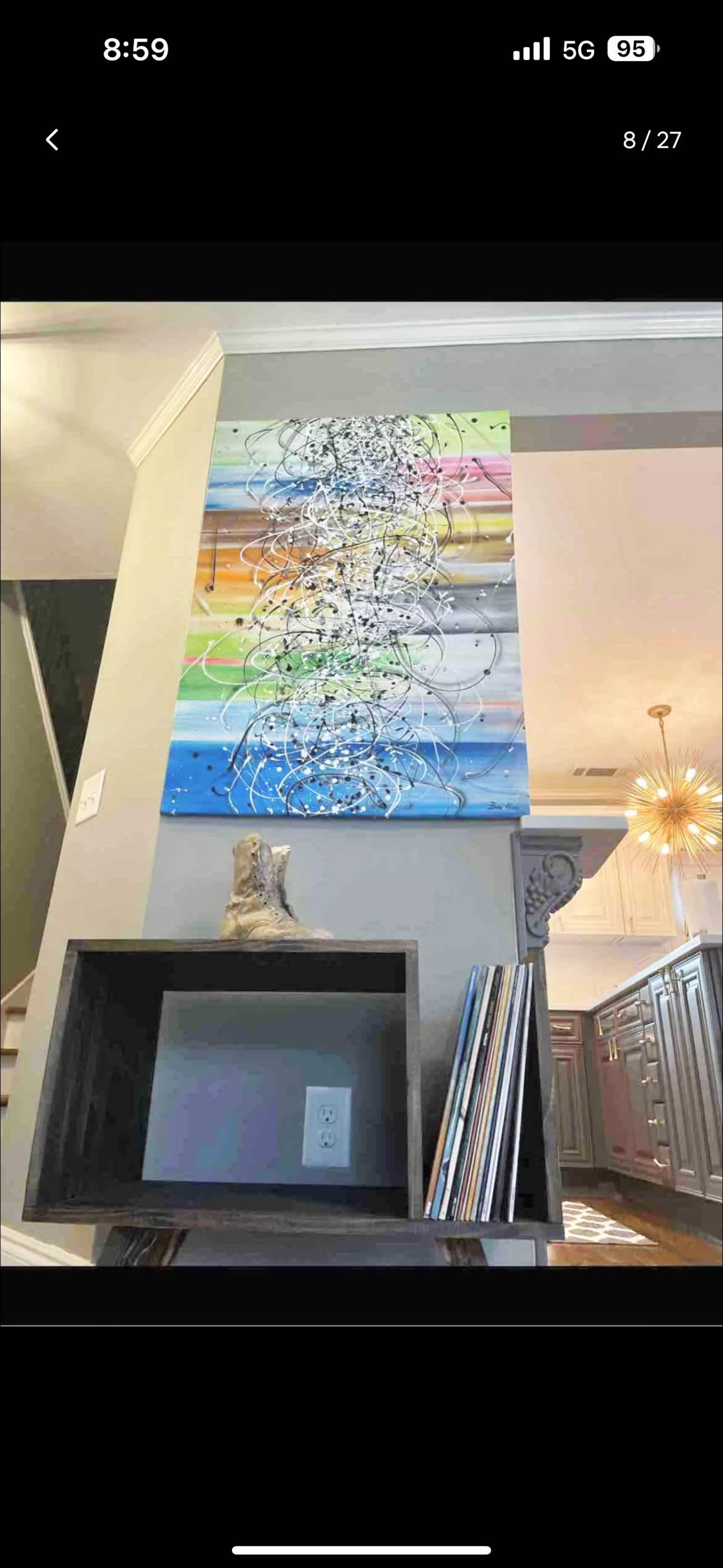 The image shows a colorful abstract painting hanging on a wall above a small shelf with records and a decorative object.