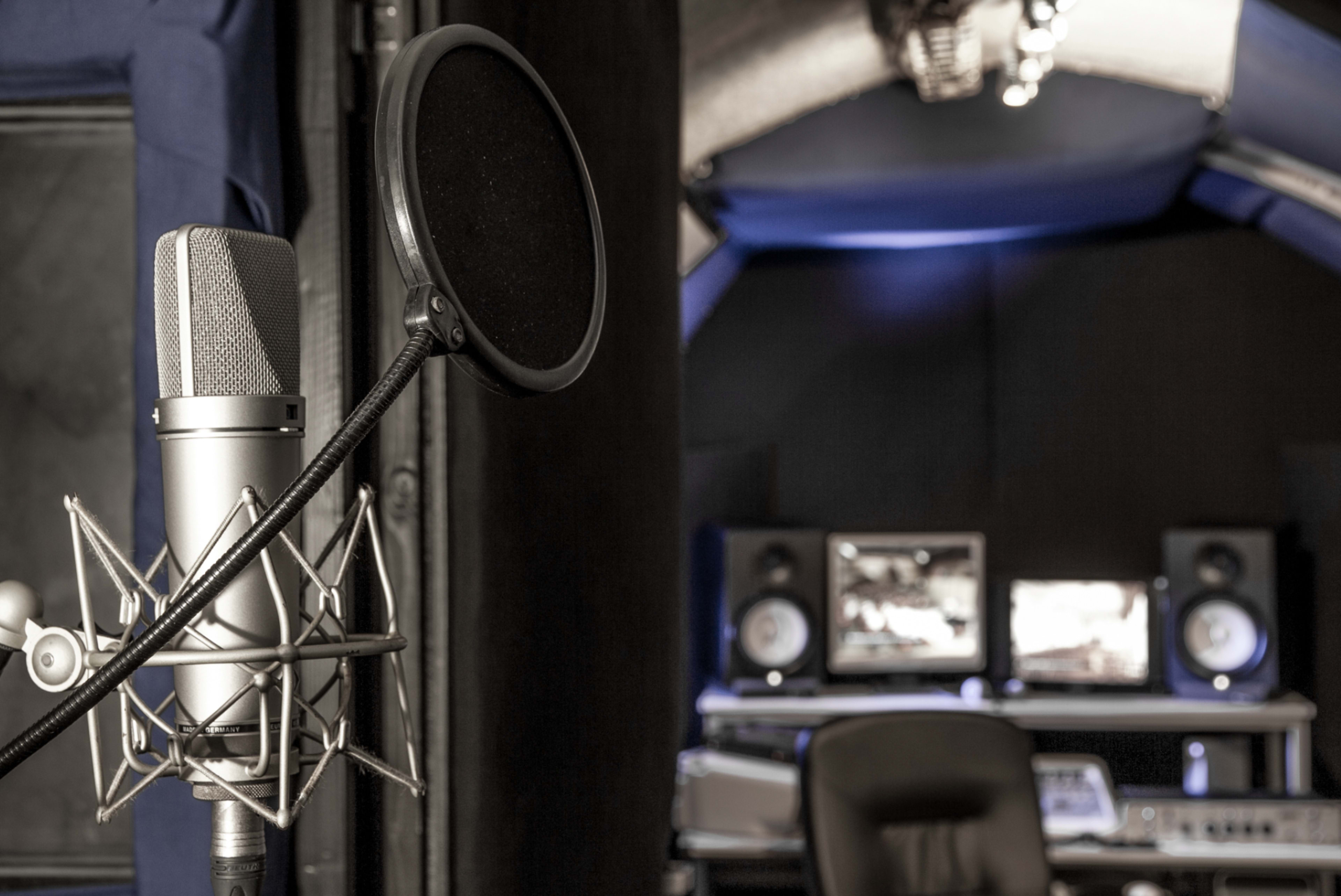 A microphone with a pop filter stands in the foreground of a sound recording studio, featuring computer monitors and audio equipment in the background.