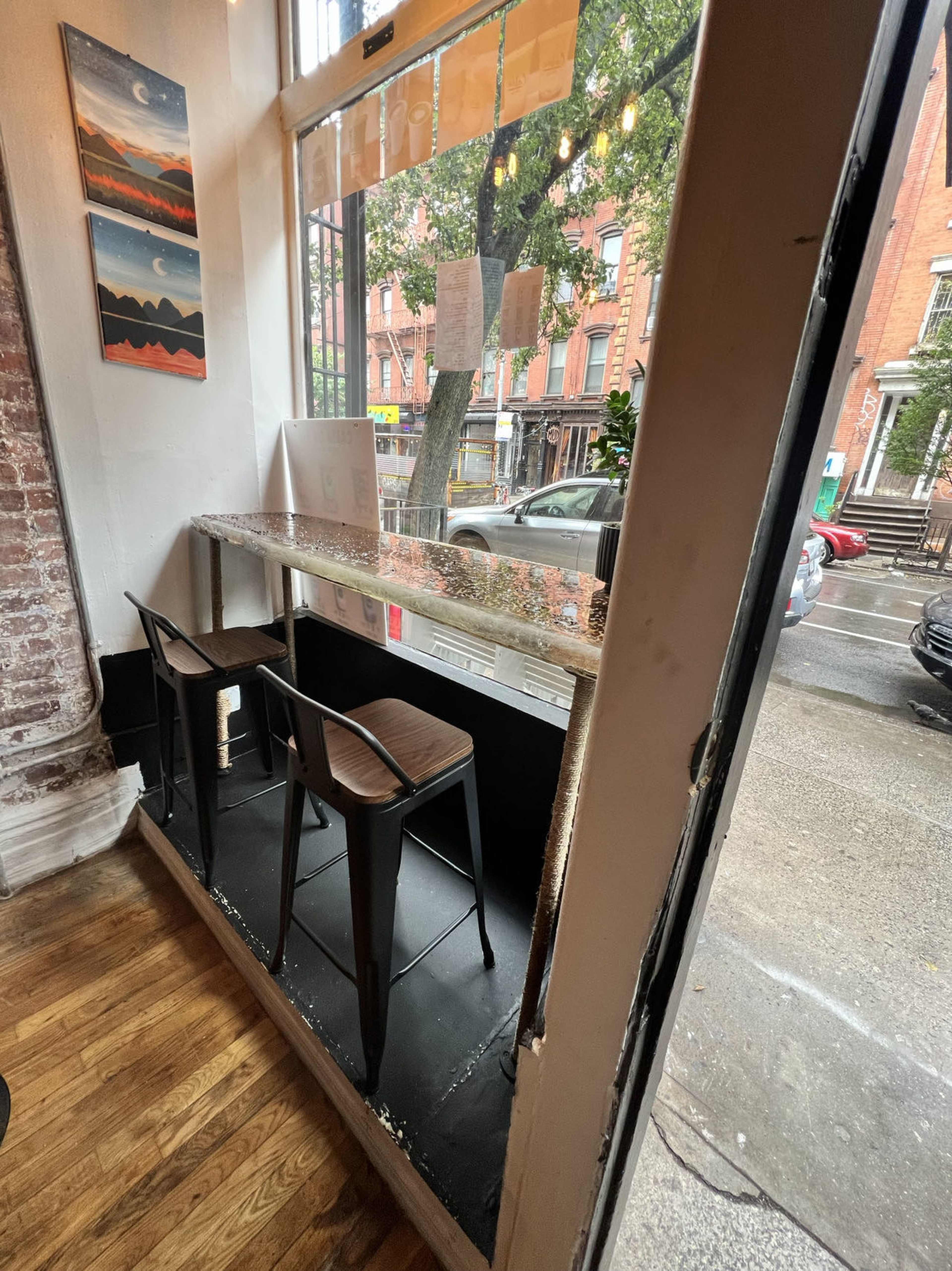A small cafe space featuring two bar stools at a window counter, with a view of a rainy street outside.