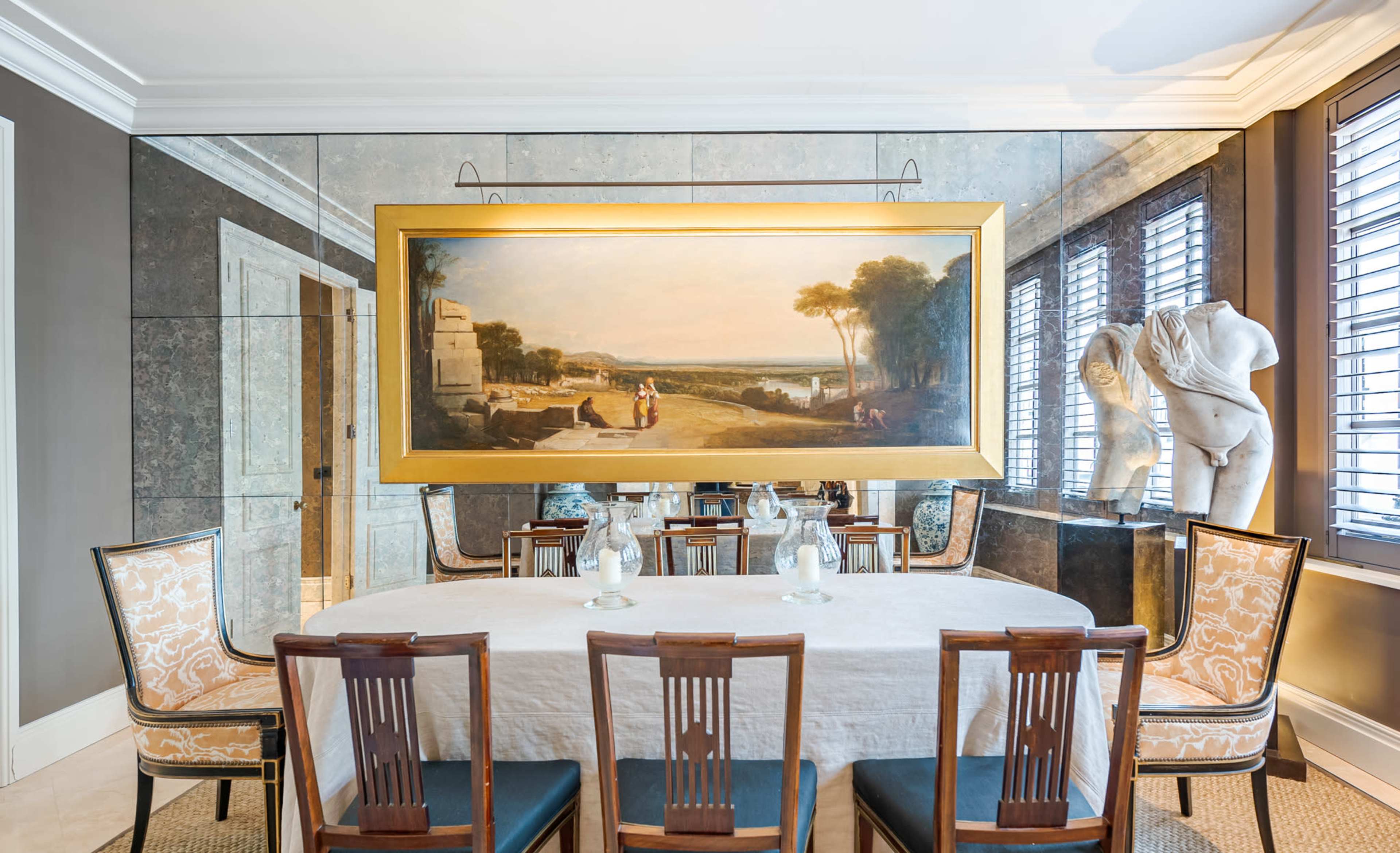 A dining room features a large painting of a landscape on the wall, with a white tablecloth-covered table surrounded by wooden chairs and a sculpture in the corner.