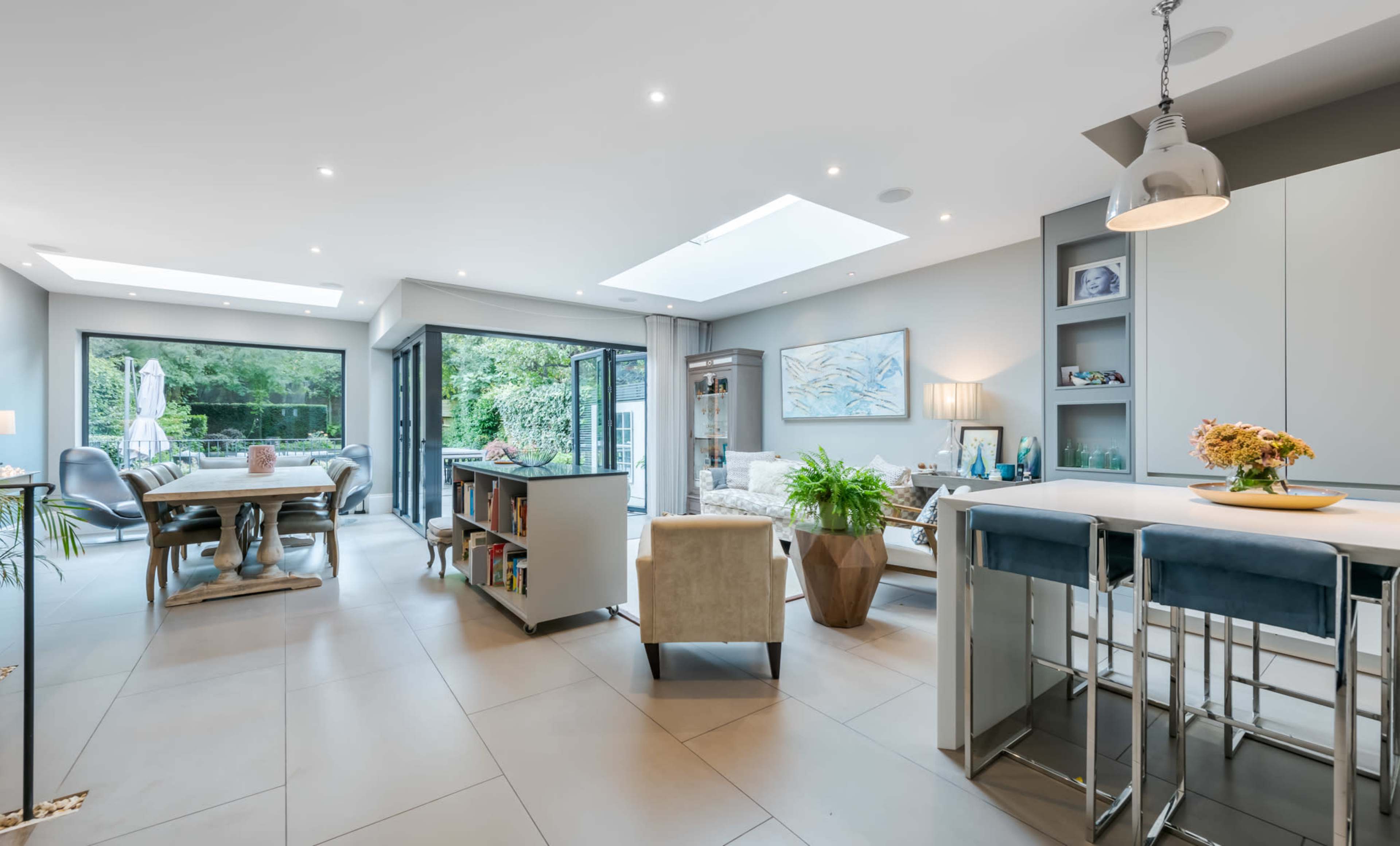 The image shows a spacious modern kitchen and living area with large glass doors opening to a garden, featuring elegant furniture and a neutral color scheme.