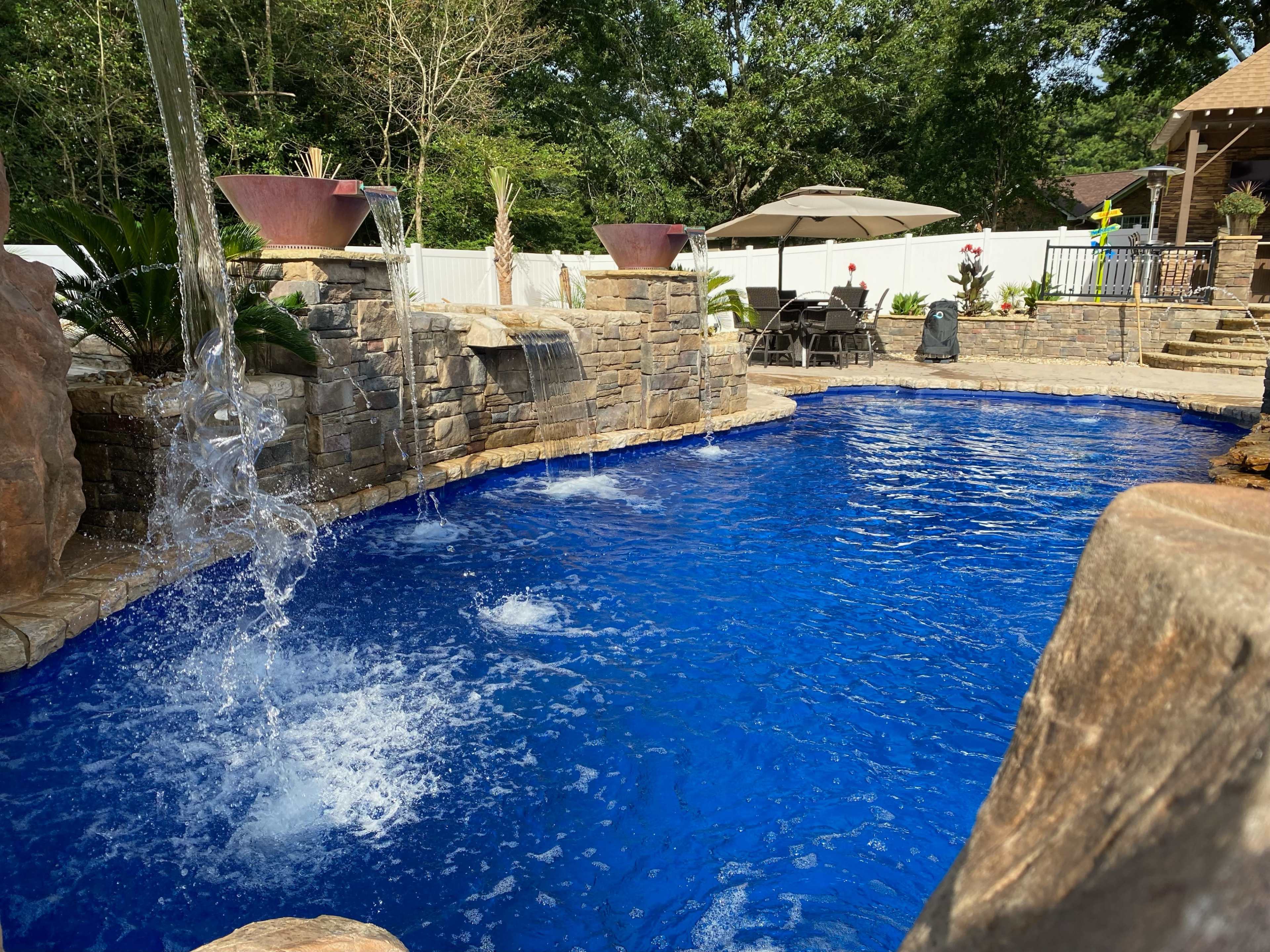 A backyard pool surrounded by stone walls, a waterfall feature, and a shaded seating area.