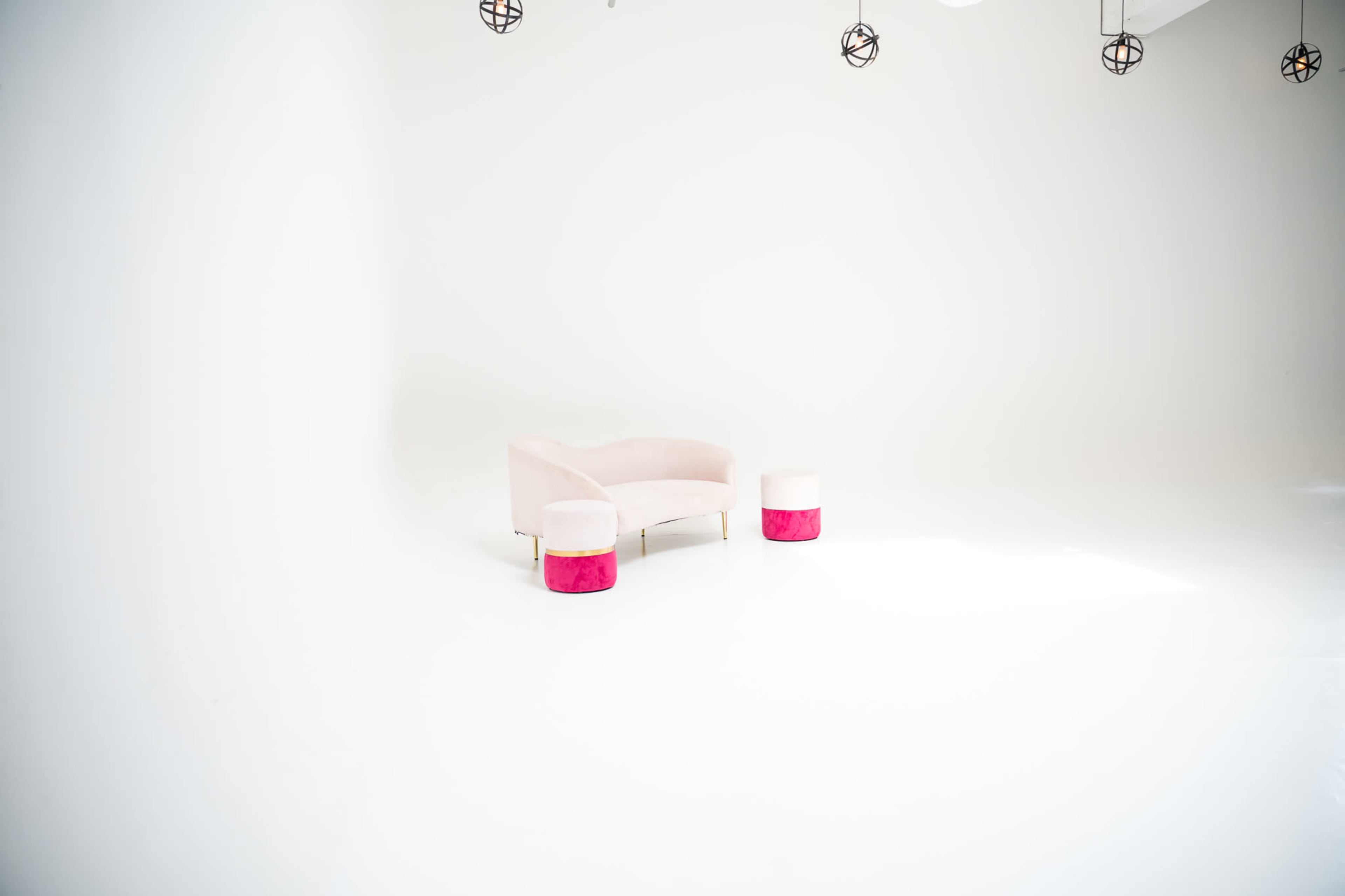 A pink sofa and matching ottoman are placed in a brightly lit, minimalistic white room with pendant lights overhead.