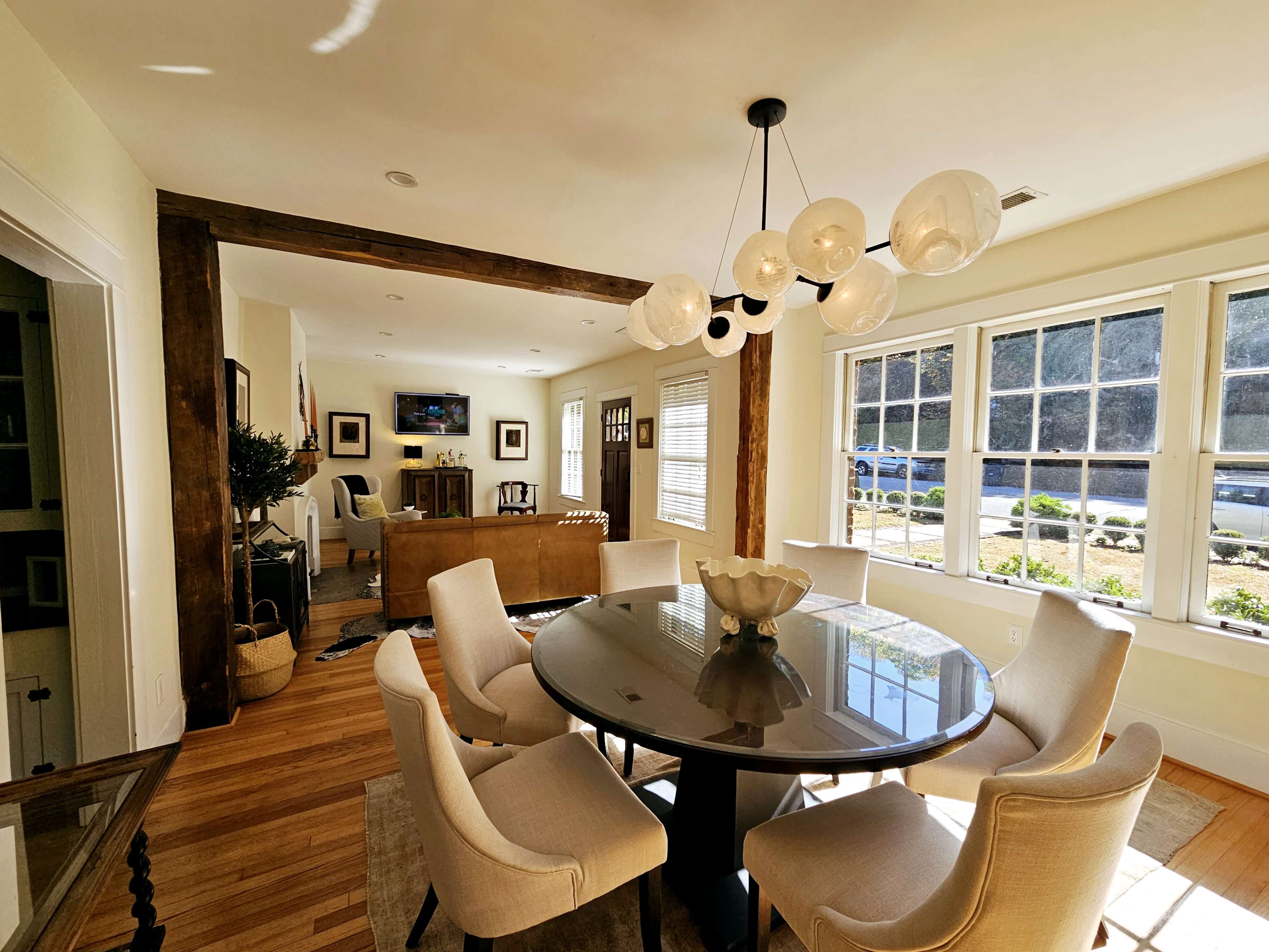 A round dining table with eight chairs is positioned in a well-lit room featuring large windows and a chandelier.