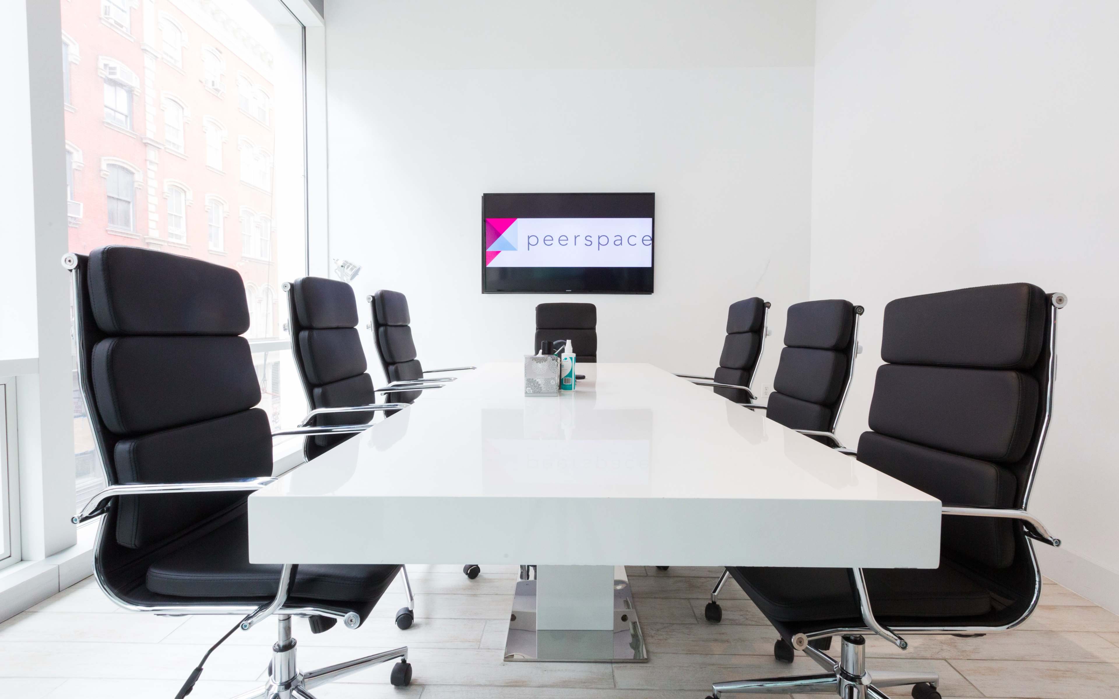 A modern conference room features a long white table surrounded by black ergonomic chairs, with a large screen displaying the Peerspace logo on the wall.