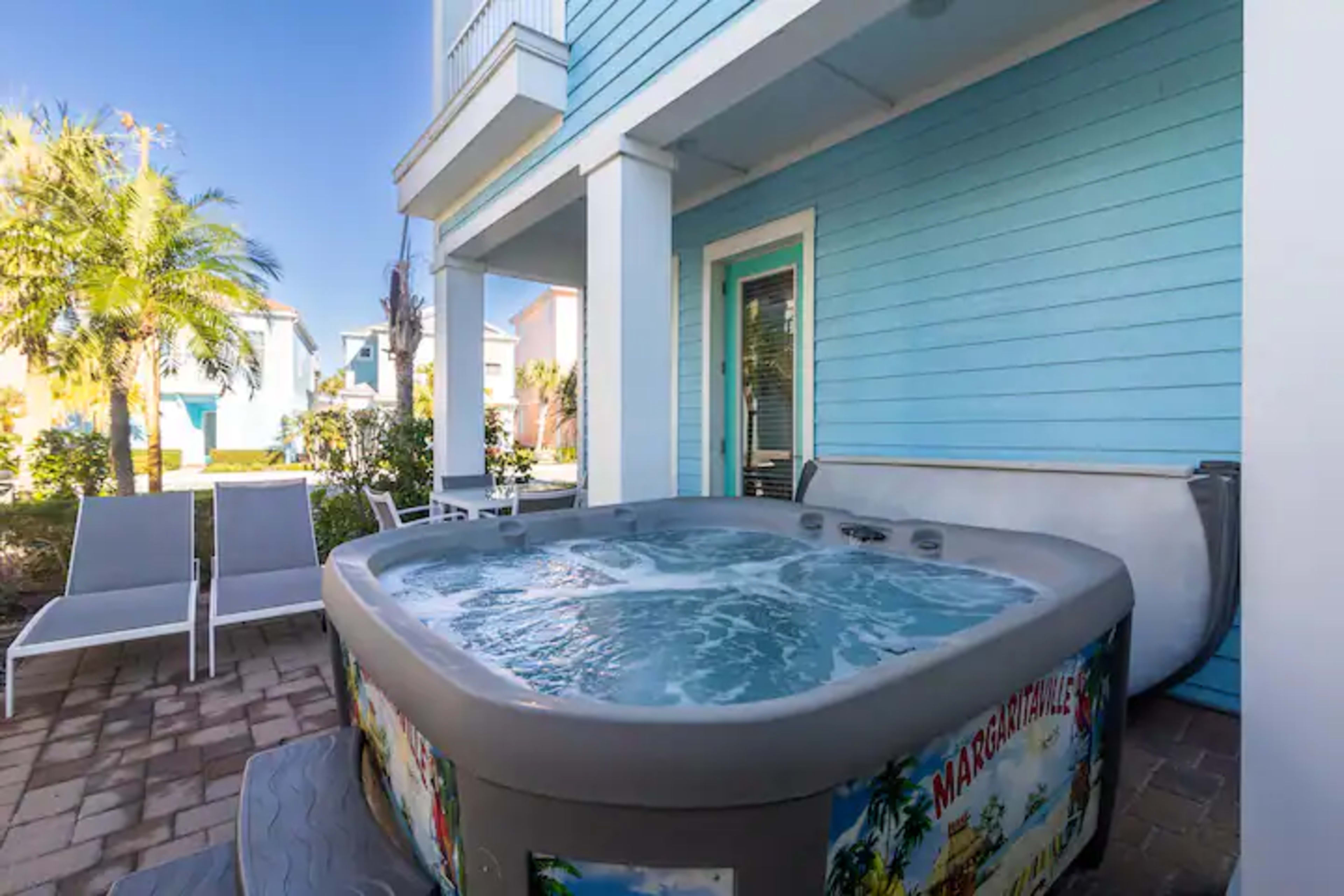 A hot tub filled with water sits beside a patio area with lounge chairs and palm trees in a residential setting.