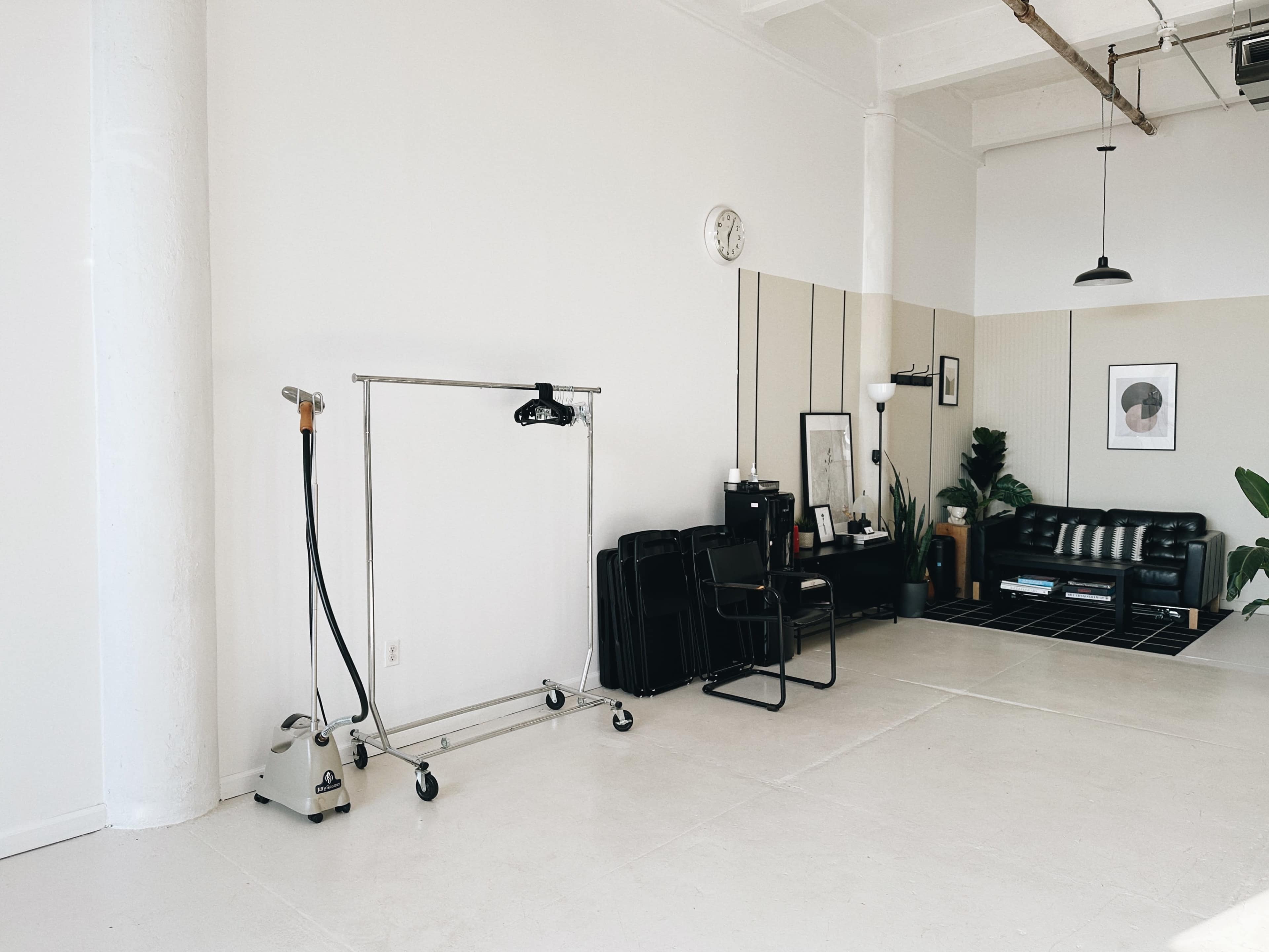 The image shows a minimalist studio space with a clothing rack, a steaming appliance, a black leather sofa, and potted plants against light-colored walls.
