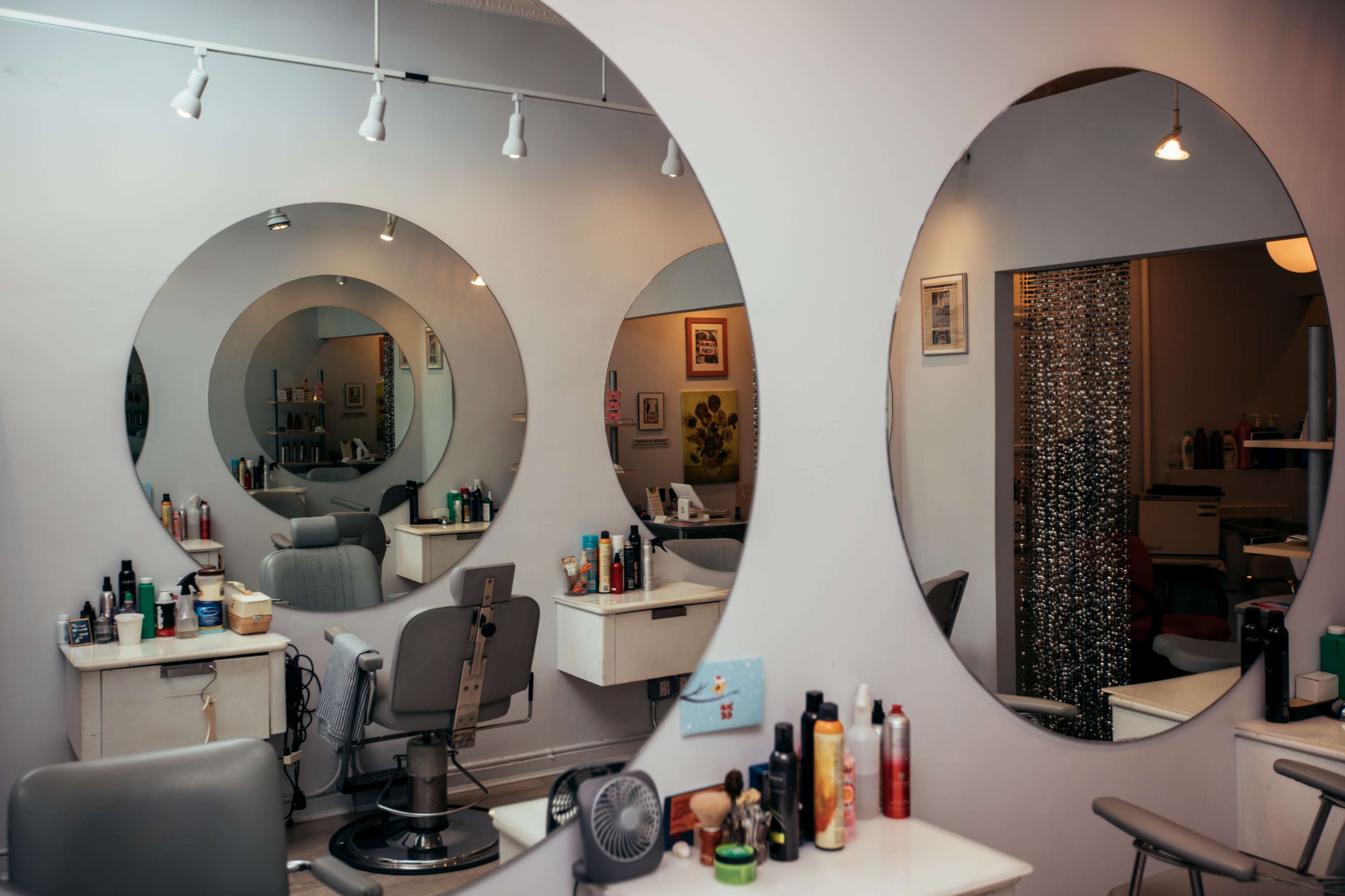The image shows a hair salon with three large circular mirrors reflecting styling chairs and various haircare products on a white wall.